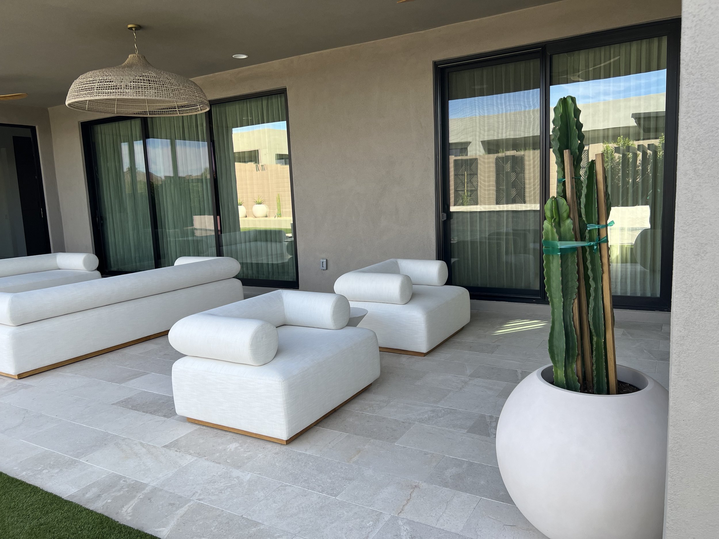 Outdoor patio area with white lounge seating, a large potted cactus, glass sliding doors with green curtains, and a beige wall.