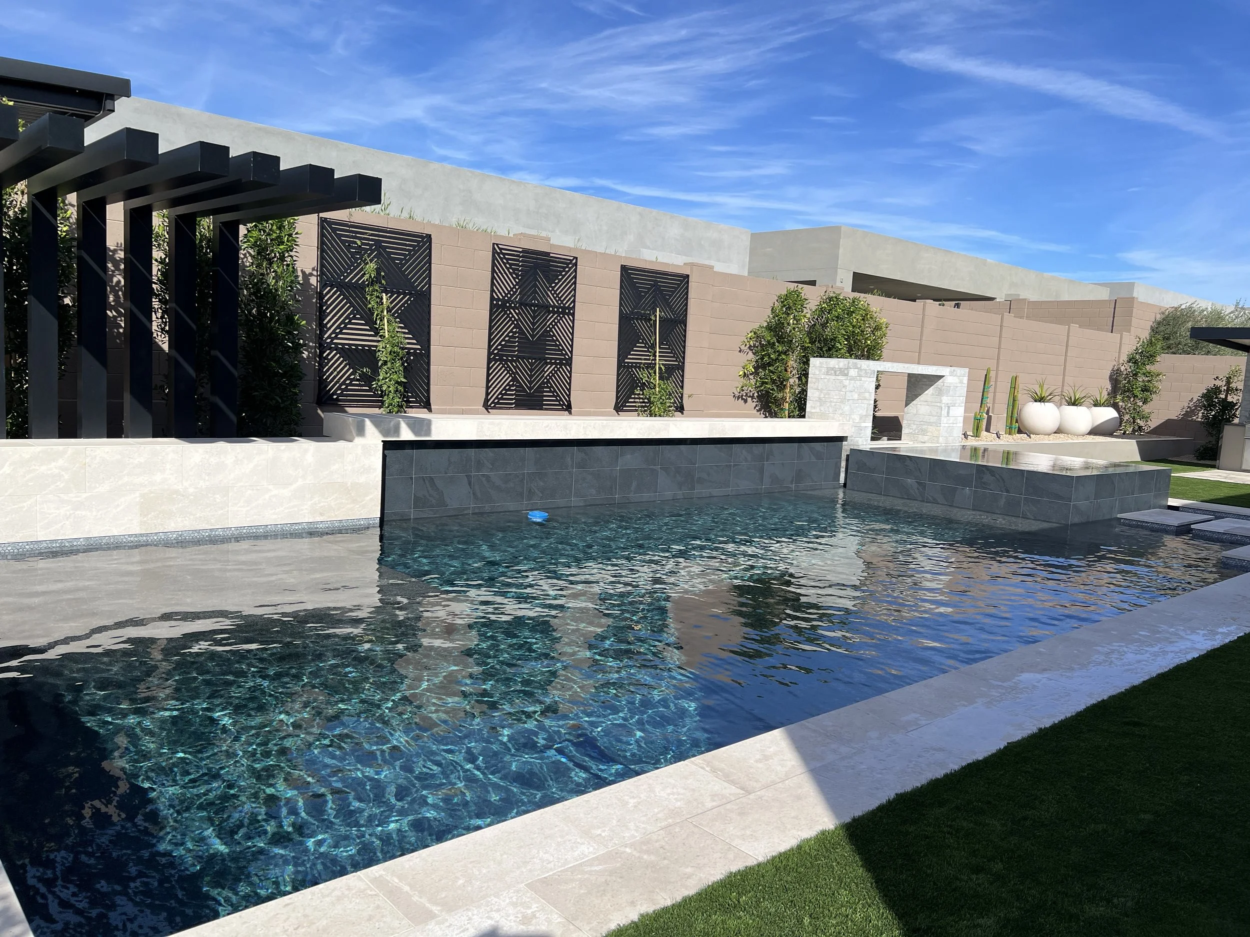 Modern backyard with a swimming pool, decorative stone wall with lattice panels, potted plants, and a clear blue sky.