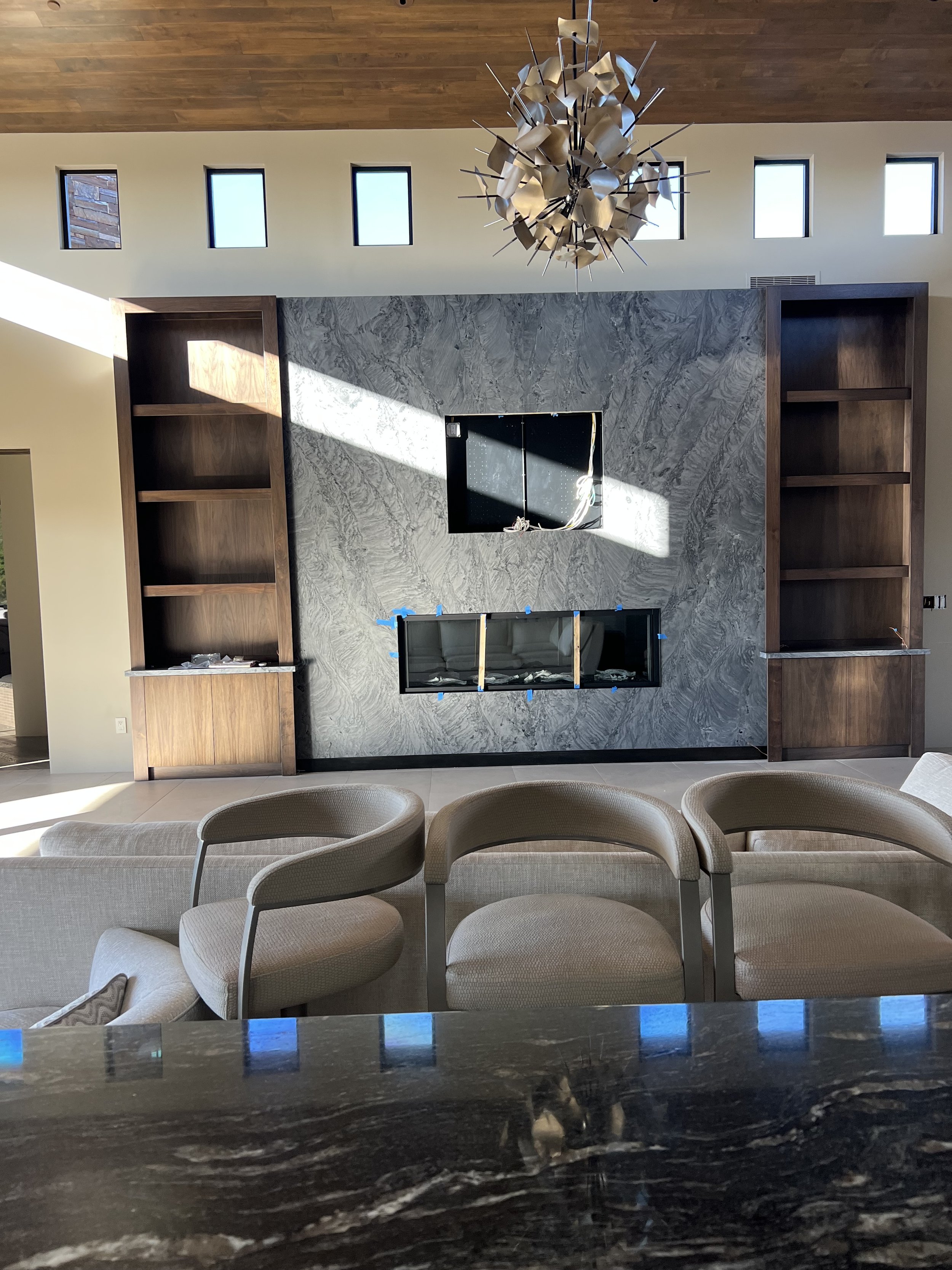 Living room with a marble fireplace surround, built-in wooden shelves, beige chairs with curved backs, and a modern chandelier hanging from a wooden ceiling. Five rectangular windows are above the fireplace, with natural light streaming in.