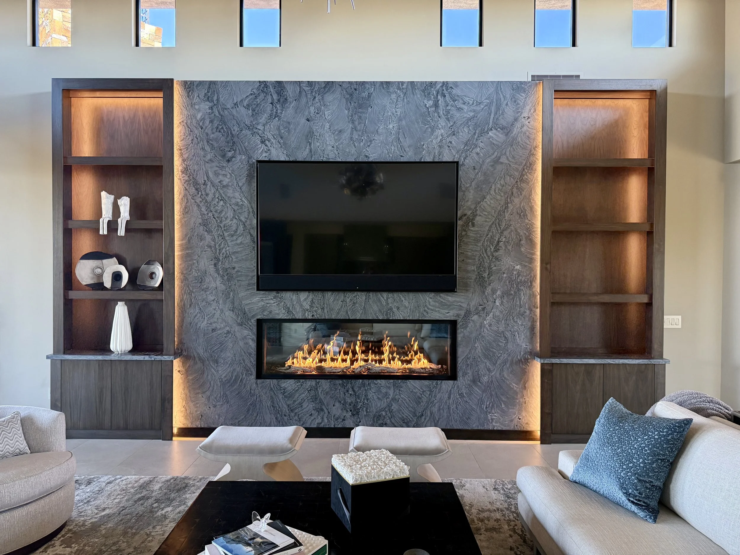 Modern living room with a marble fireplace surround, a flat-screen TV, wooden shelving, and beige sofas with throw pillows.