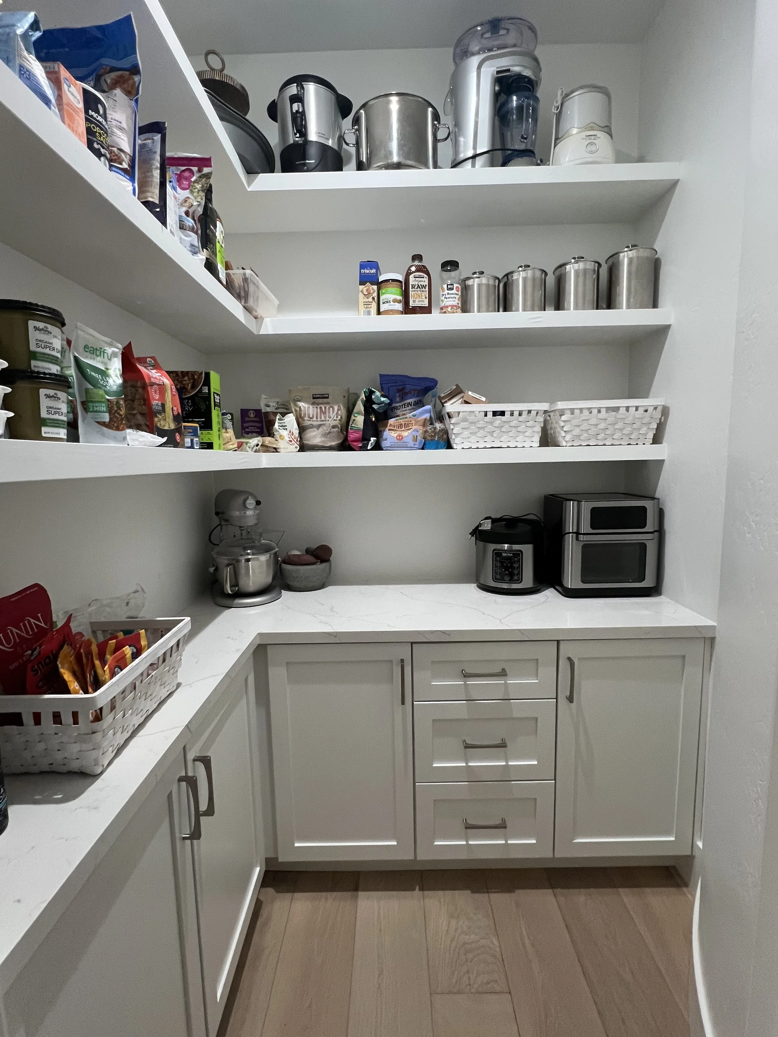 A kitchen pantry with three white shelves filled with various food items, kitchen appliances, and storage containers, with white cabinets and wooden flooring.