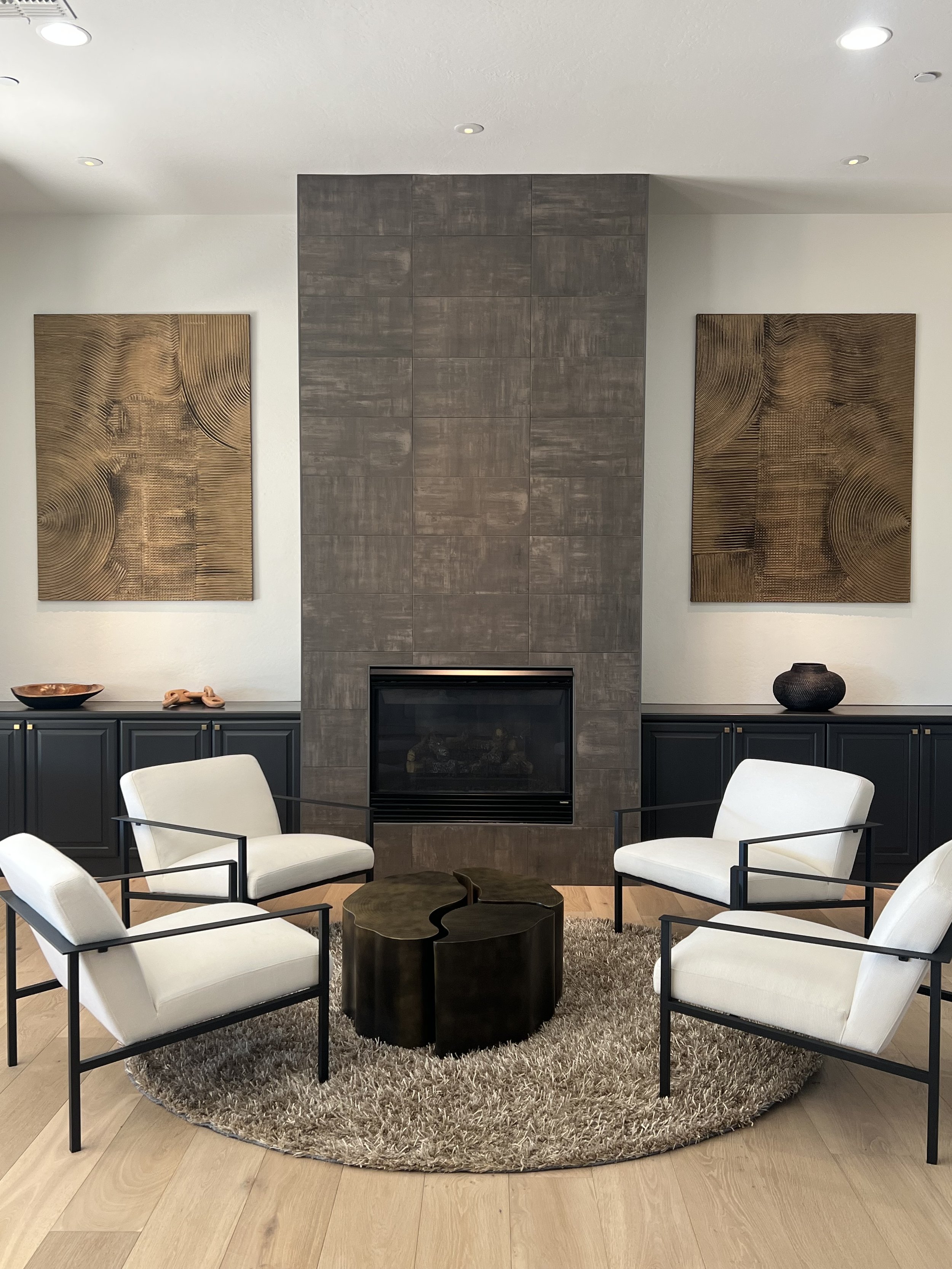 Living room with a fireplace, two abstract wooden wall art pieces, four white armchairs with black frames, a round wooden coffee table, a shaggy rug, and a cabinet with decorative items.