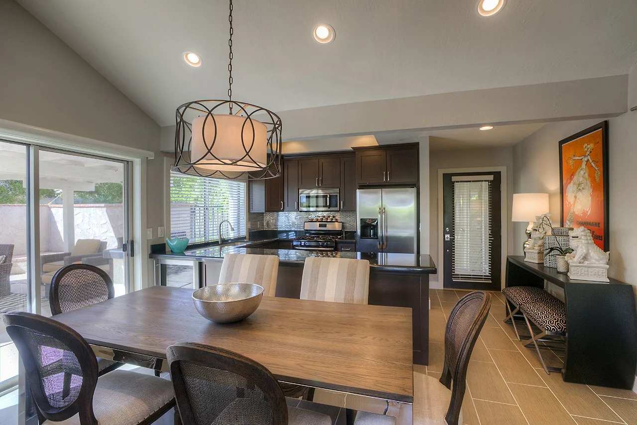 Kitchen and dining area with wooden table, six chairs, dark cabinets, stainless steel appliances, sliding glass door, window with blinds, and decorative art piece.