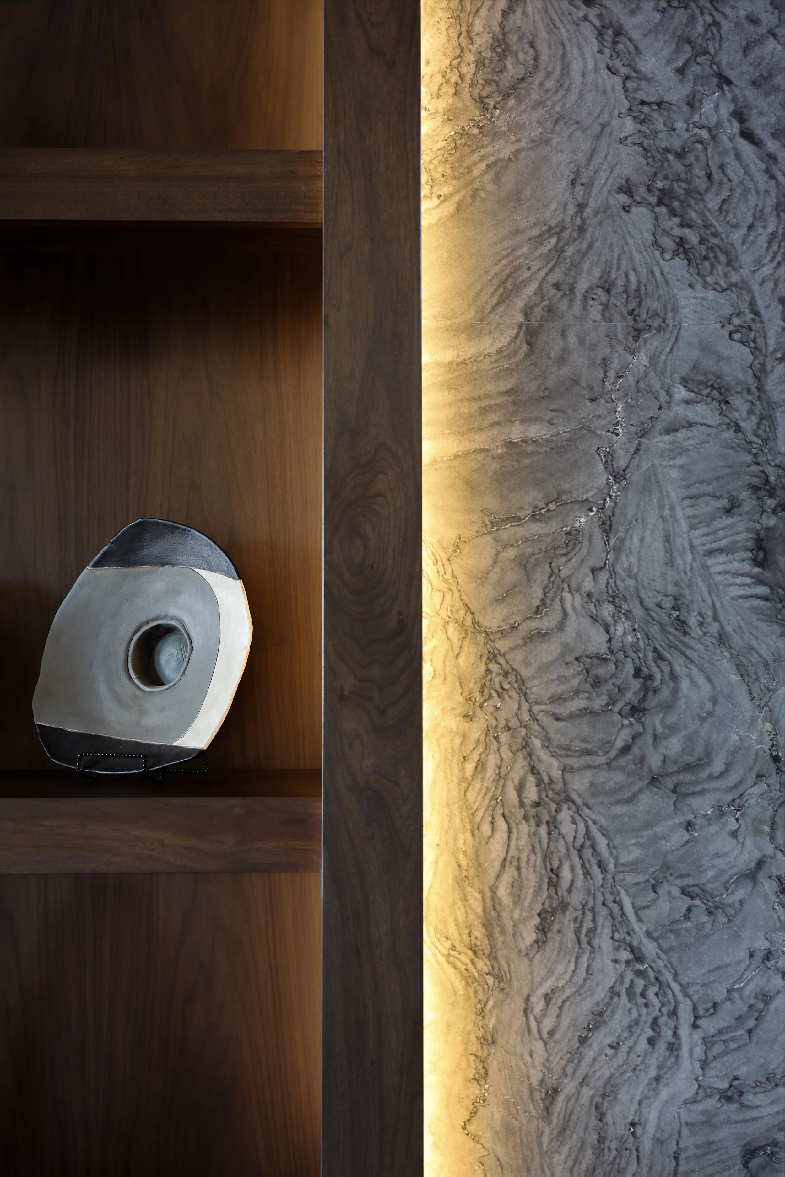 A close-up photo of a wooden bookshelf with a carved wooden mask placed on it, next to a wall with marble design and warm lighting.