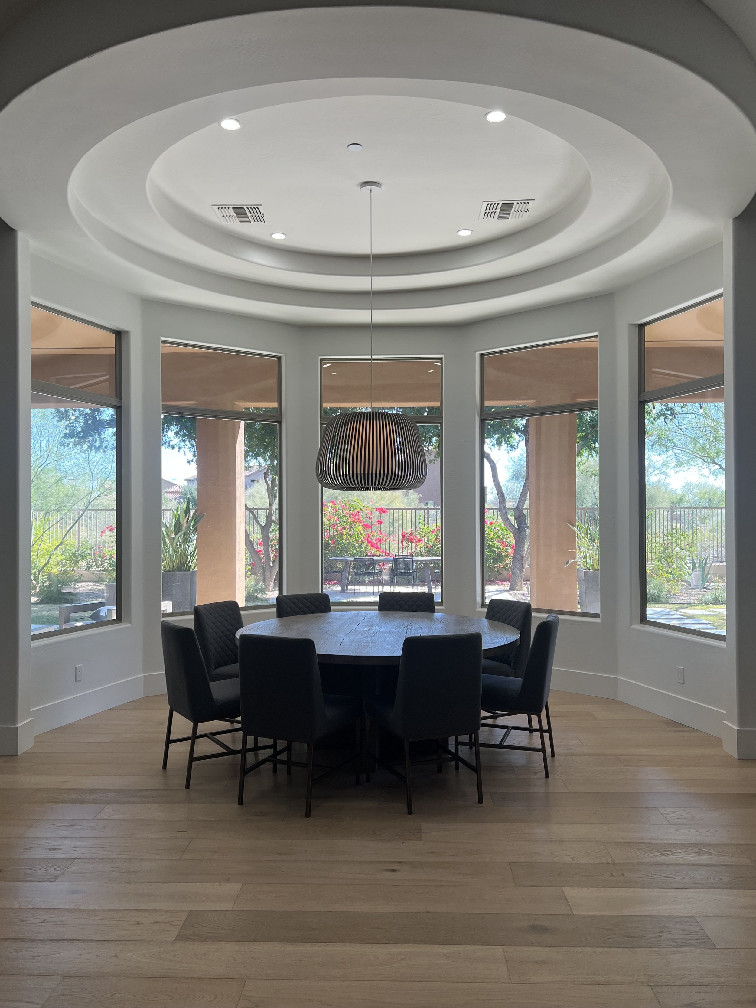 A round dining table with six black chairs inside a bright room with large windows overlooking a garden. There is a modern black pendant light hanging above the table. The room has light-colored wooden floors and white walls with a circular ceiling d