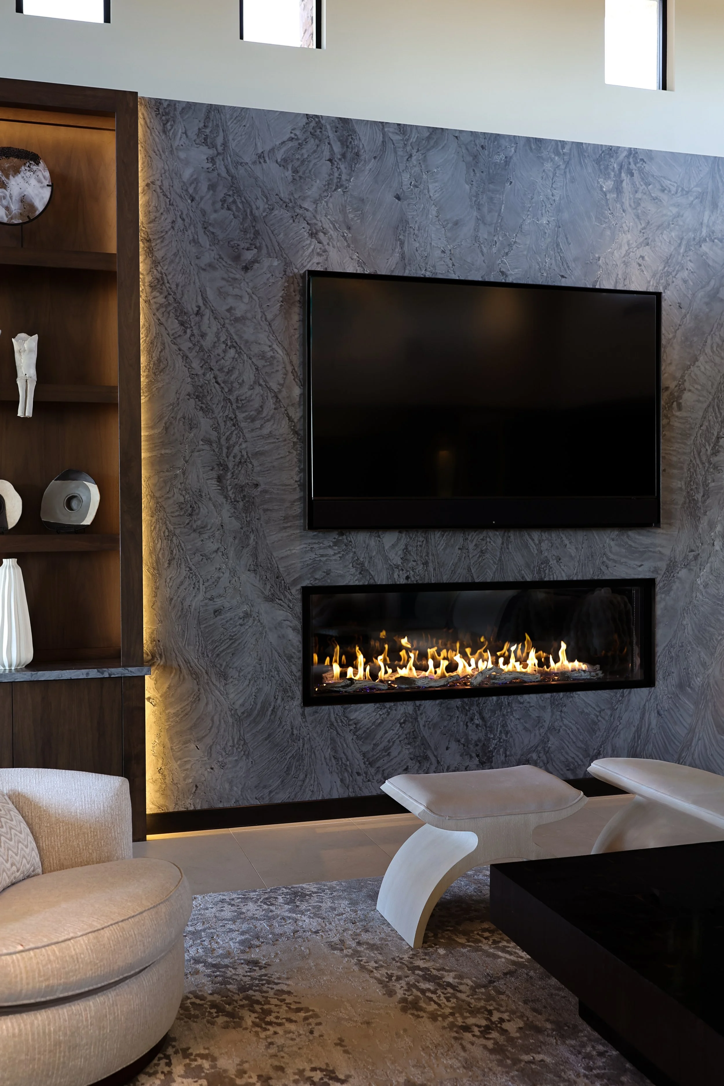 Modern living room with a marble wall, wall-mounted flat-screen TV, and a fireplace with flames. There is a beige armchair and a white modern lounge chair on a patterned rug, with three small windows high on the wall.