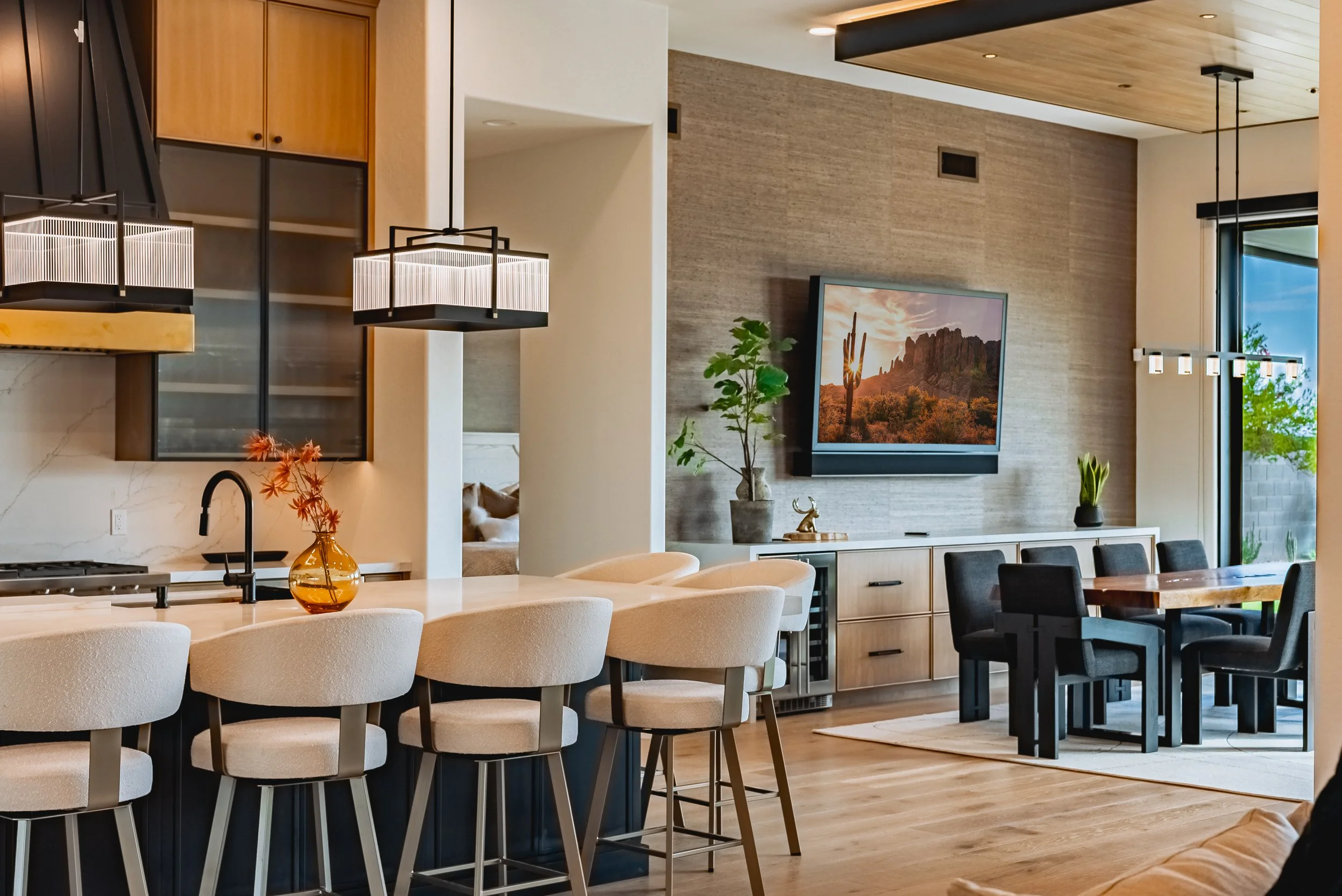 Modern open-concept living and dining area with wooden accents, a large framed landscape on the wall, potted plants, and contemporary lighting fixtures.