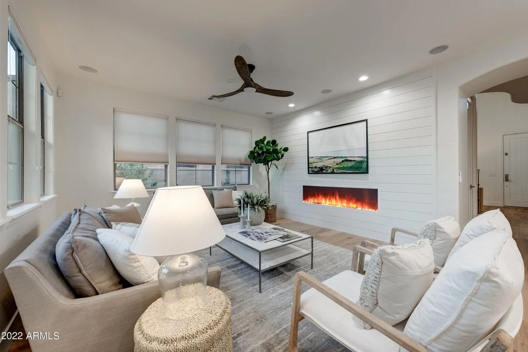 Living room with beige sofas, white armchairs, a modern fireplace, a large screen TV, large windows with shades, a ceiling fan, and a potted plant.