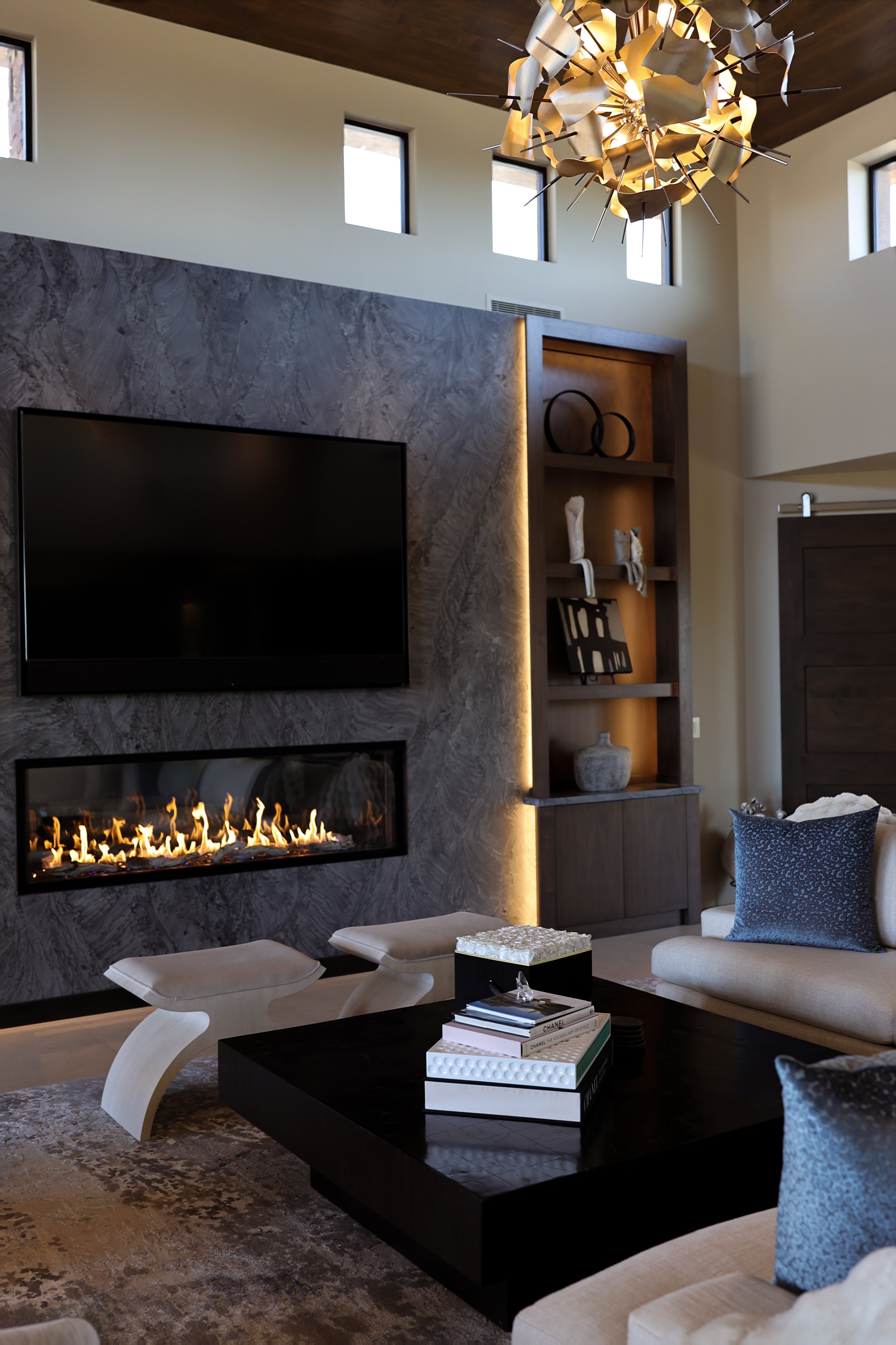 Modern living room with a wall-mounted fireplace, a black coffee table with books, a beige sofa with blue pillows, decorative shelves with sculptures and art pieces, and a stylish chandelier.
