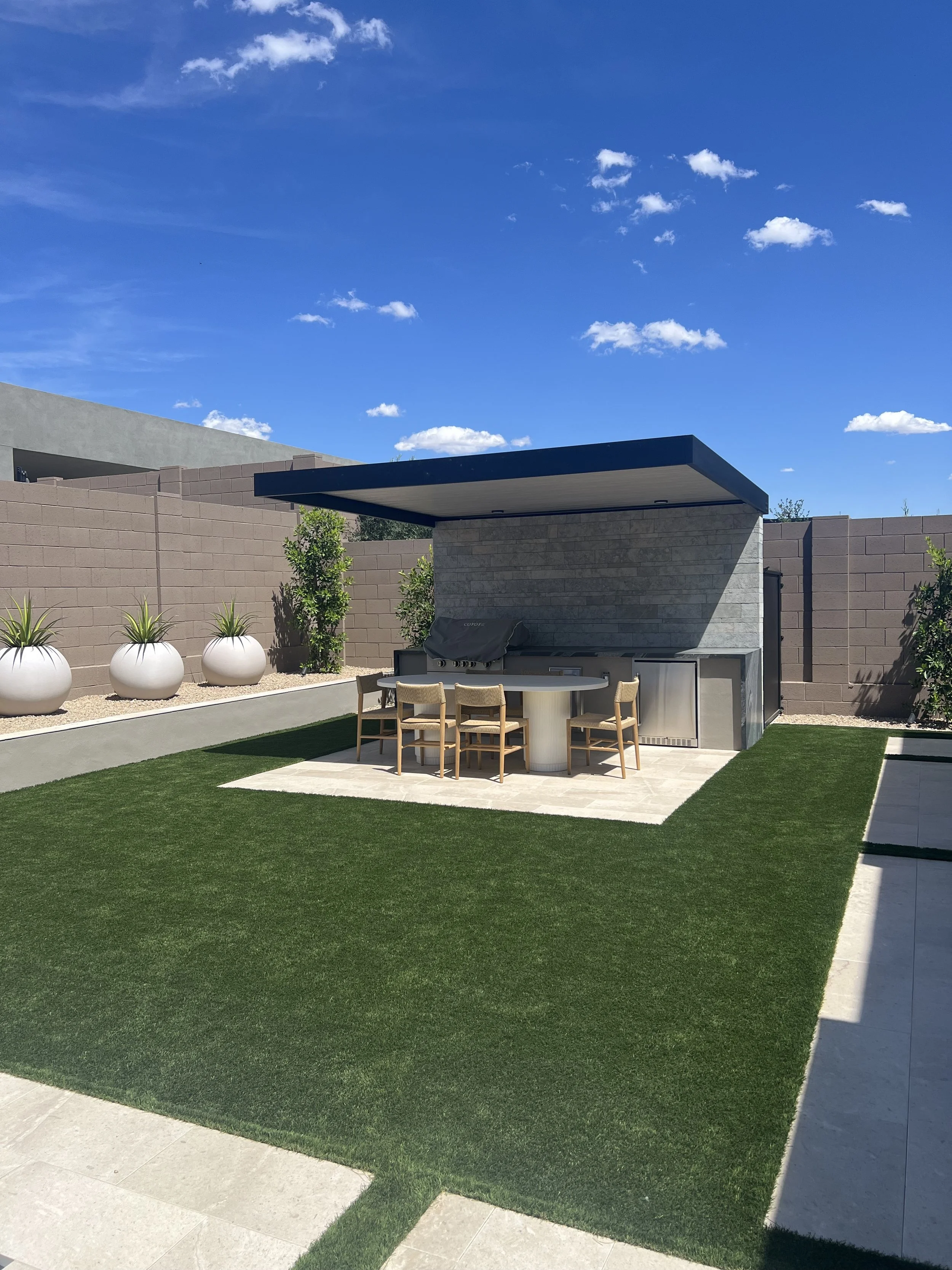 A modern backyard with a green artificial grass lawn, a concrete patio, a beige brick wall with potted plants, and a covered outdoor kitchen area.