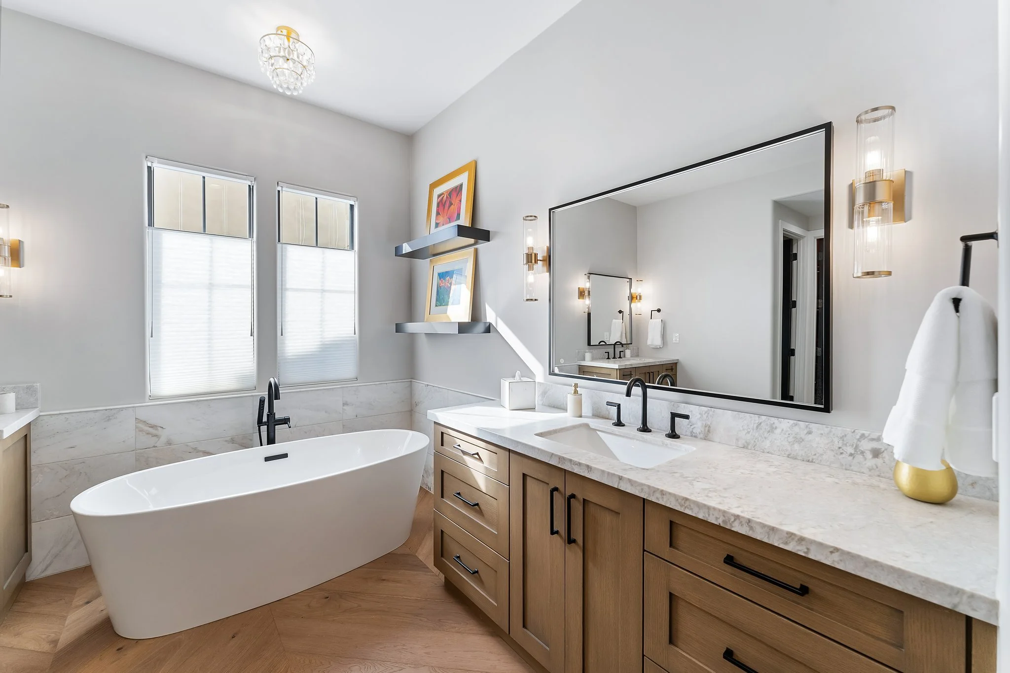 Modern bathroom featuring a freestanding bathtub, large mirror, wooden vanity with black handles, two windows with blinds, decorative artwork, and stylish lighting fixtures.