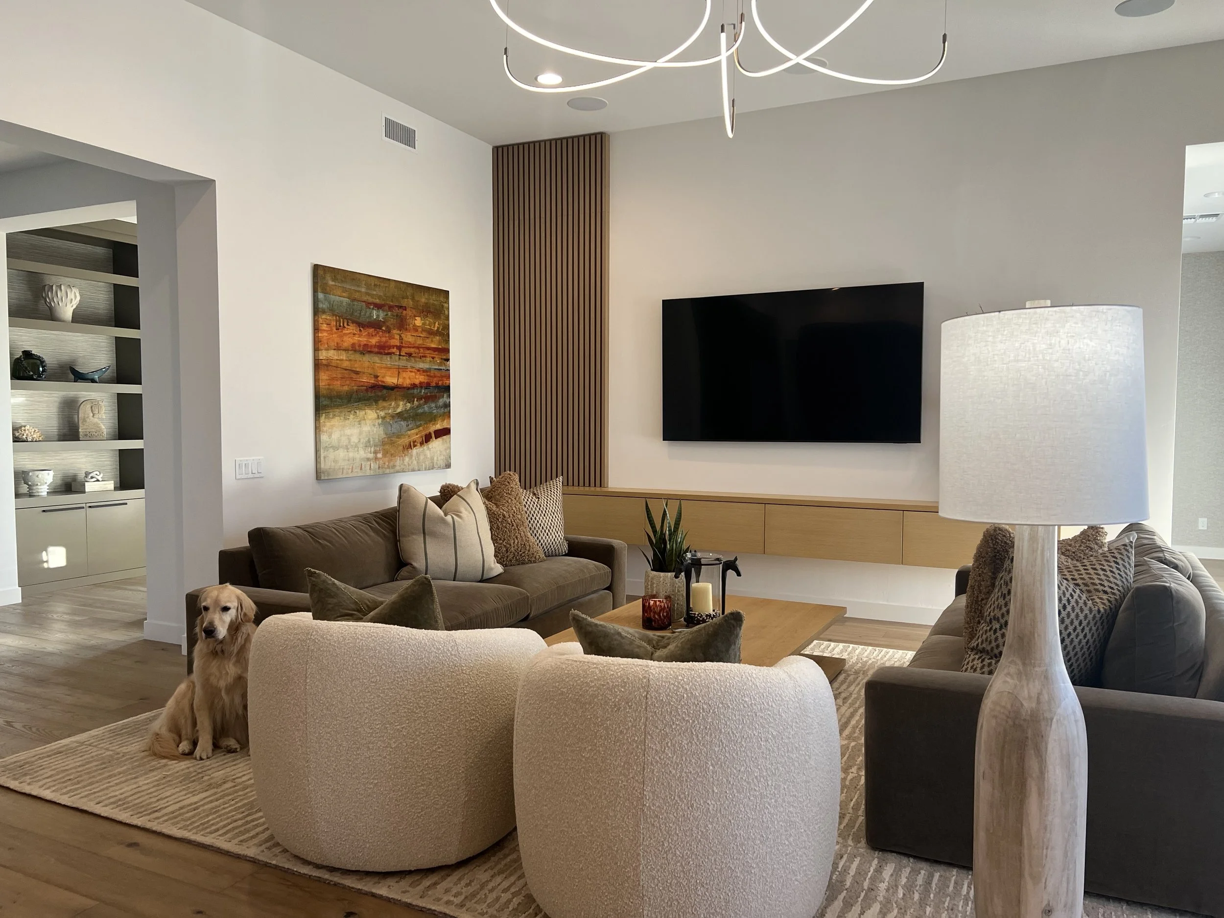 A modern living room with white walls, a wooden slatted accent wall, mounted flat-screen television, beige and gray sofas with pillows, a floor lamp, a wooden coffee table with candles and plants, and a built-in shelf displaying decorative items. A g