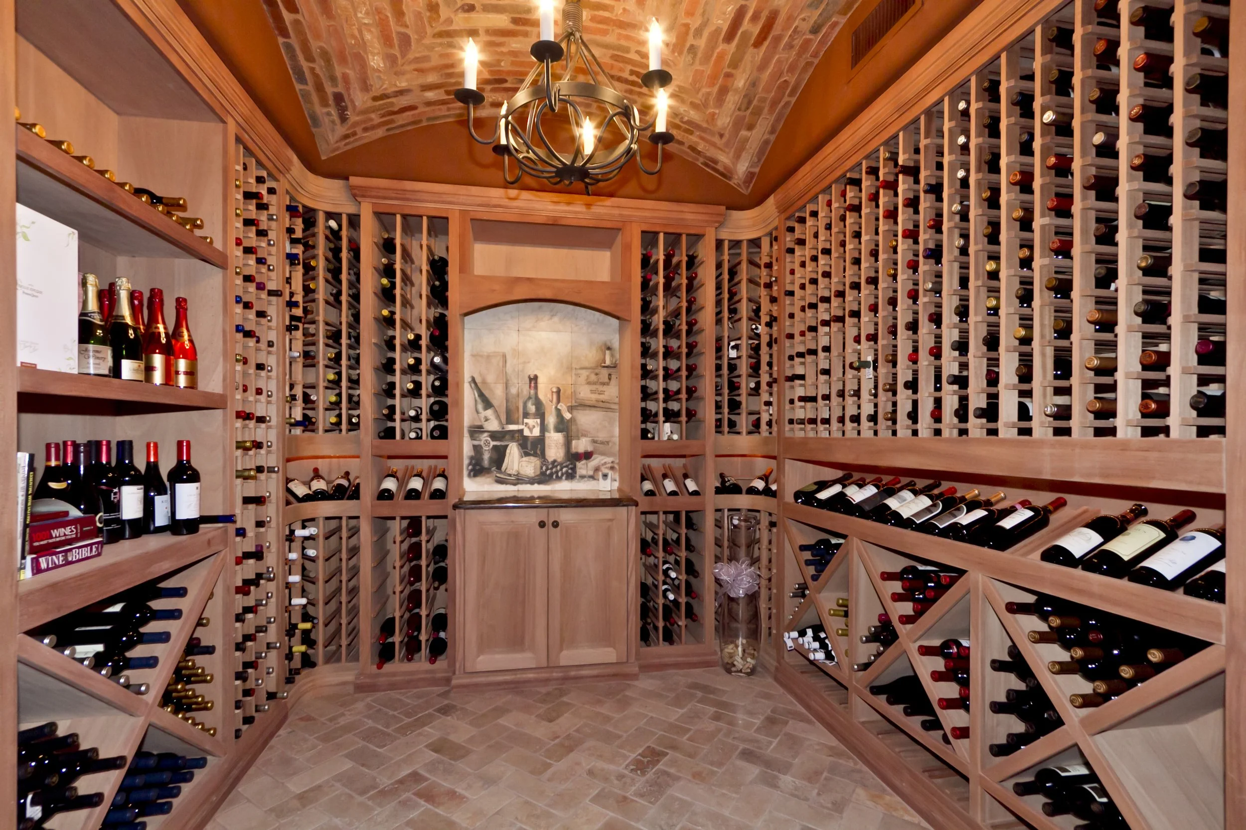 Wooden wine cellar with shelves filled with various bottles of wine, a brick vaulted ceiling, and a chandelier with candles.