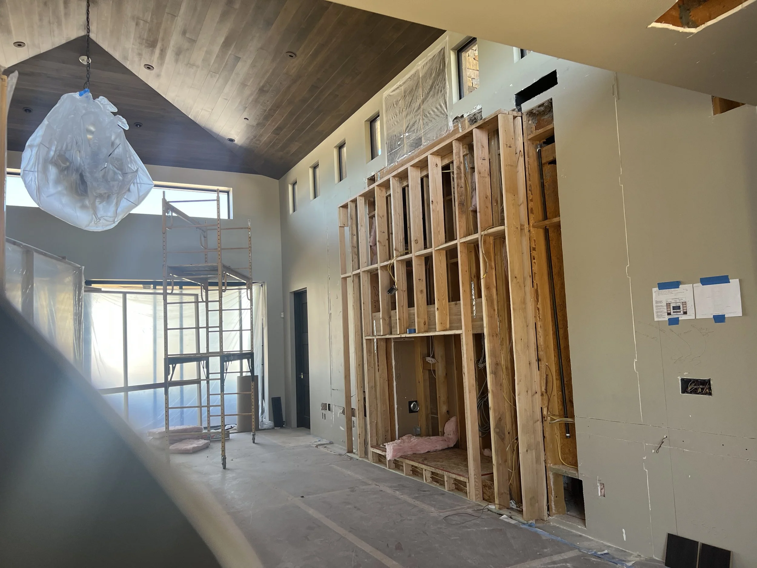 Interior of a building under construction with exposed wooden framing, drywall partially installed, and scaffolding, high windows, and construction materials visible.