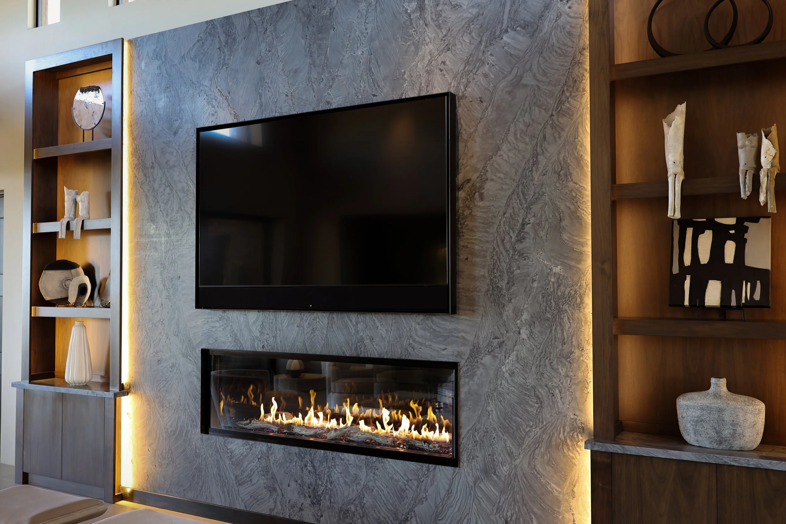 Modern living room with a mounted flat-screen TV, a marble-textured wall, built-in wooden shelves with decorative items including vases and sculptures, and a fireplace with flames.