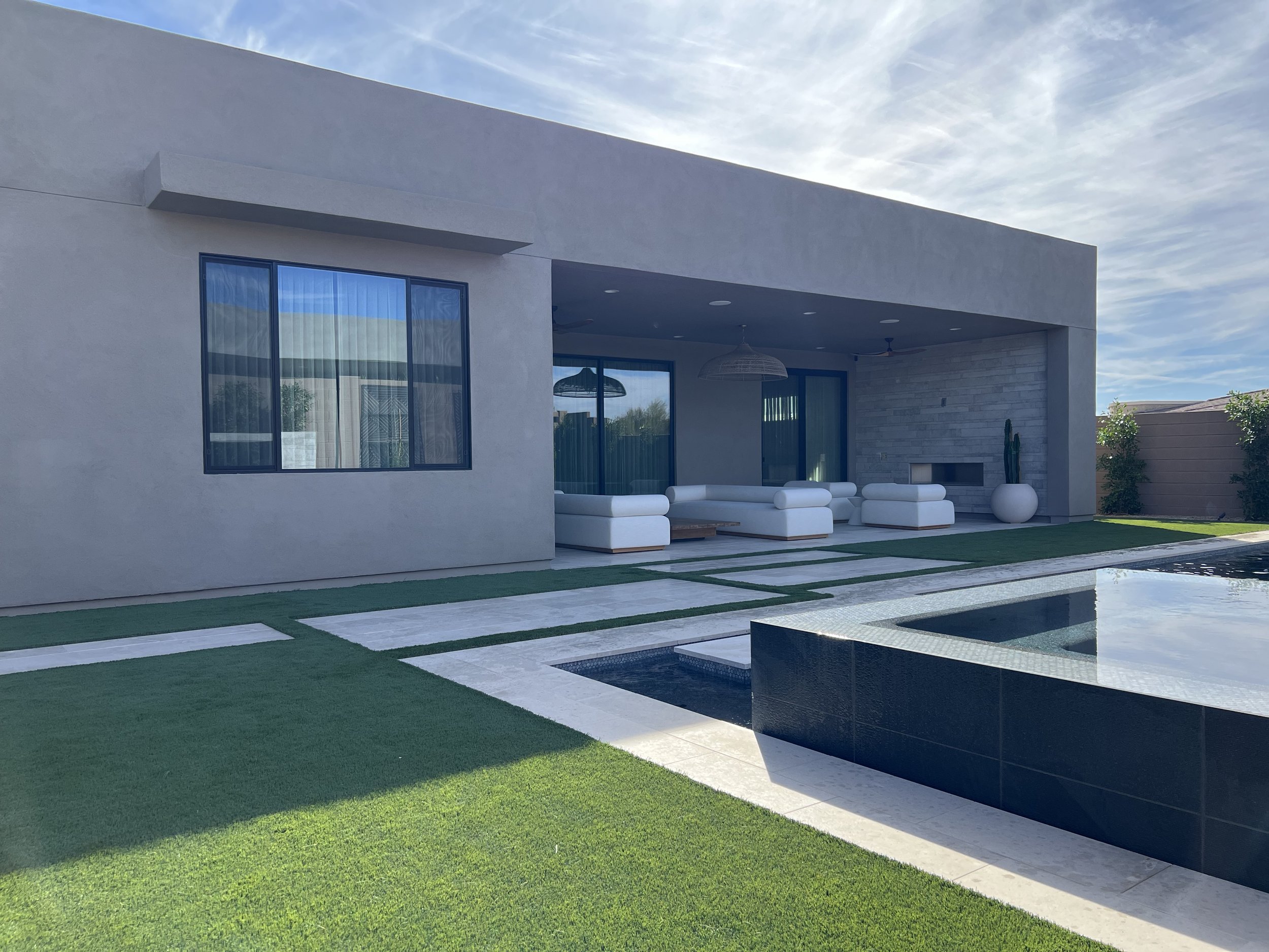 Modern house with white exterior, large glass windows, outdoor seating area with white chairs and a round table, green lawn with geometric tiles, and a pool, under a partly cloudy sky.