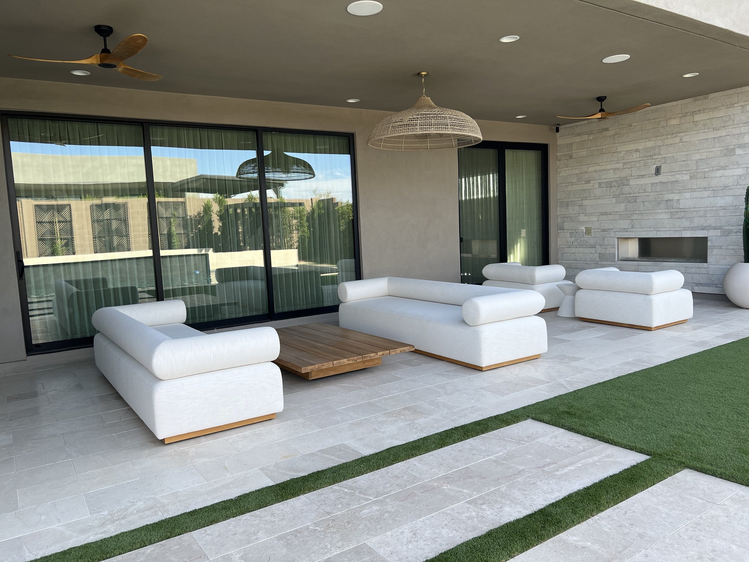 Modern outdoor patio with white cushioned seating, wooden coffee table, large glass sliding doors, stone fireplace, wicker hanging light fixture, ceiling fans, and artificial grass.