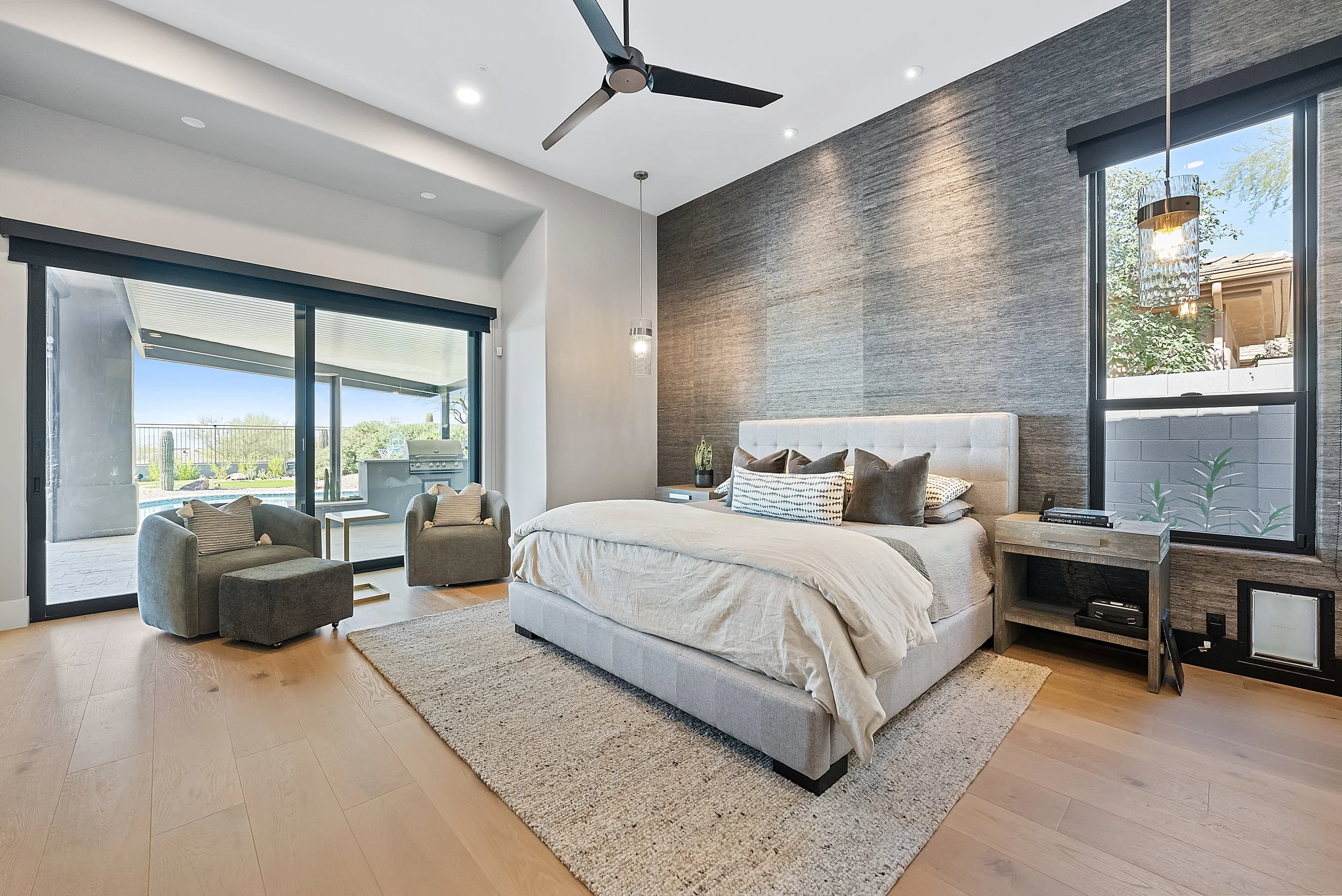 Modern bedroom with large windows showing an outdoor patio, a king-sized bed with neutral bedding, a nightstand with books and decor, ceiling fan, pendant lights, and beige area rug on light wood flooring.