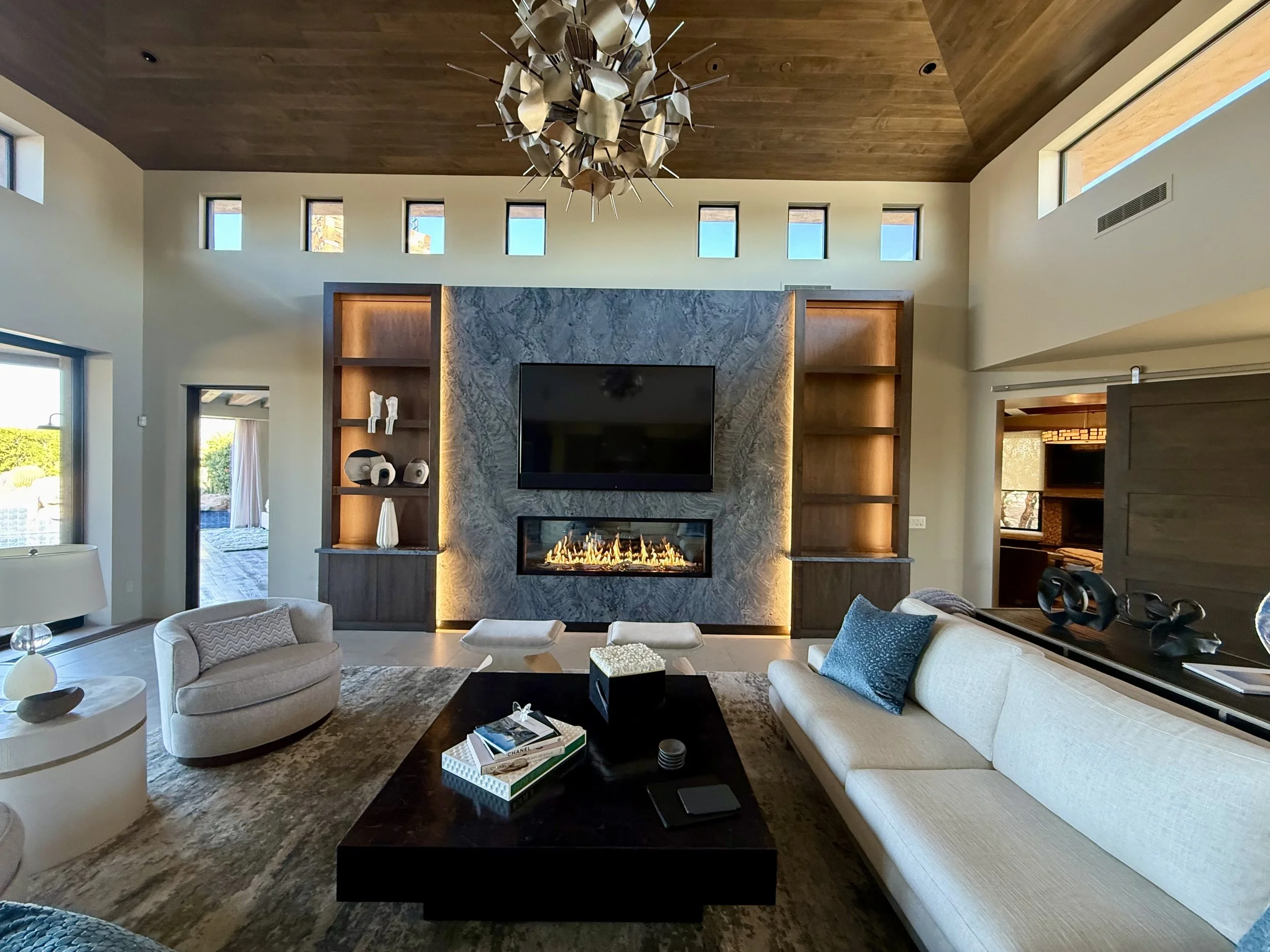 Modern living room with a fireplace, cream-colored sofa, armchair, black coffee table, built-in wood shelving, and a wooden ceiling. Multiple small windows and a chandelier are visible.