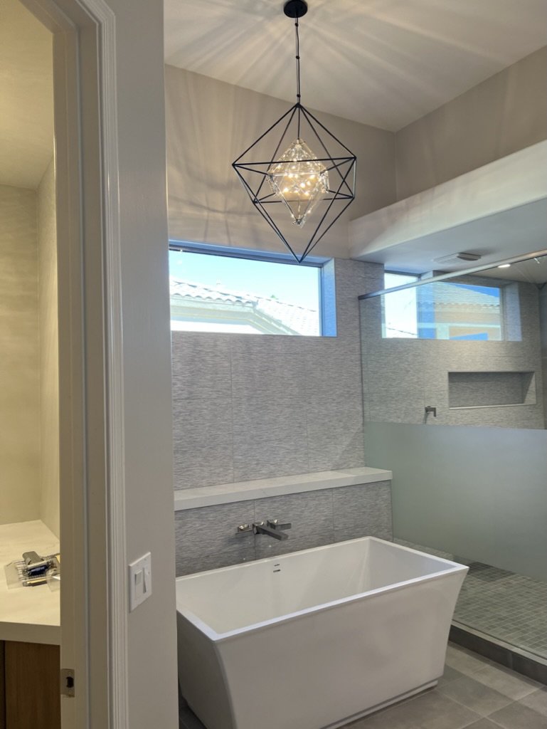 Modern bathroom with a white freestanding bathtub, gray tiled wall, a large window, and a geometric pendant light hanging from the ceiling.