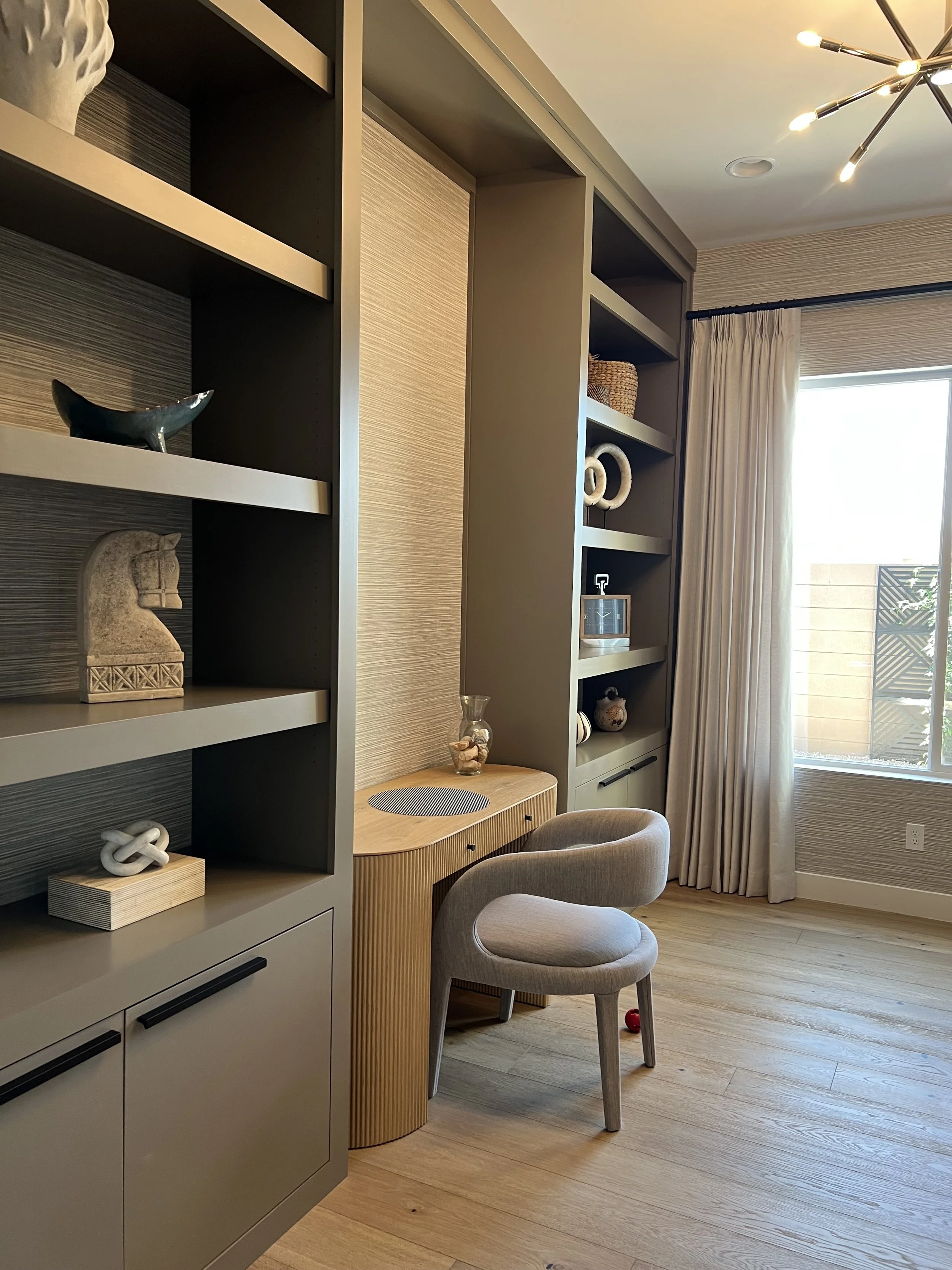 Interior view of a room with built-in shelves containing decorative items, a small wooden desk with a glass bowl, a beige upholstered chair, a large window with curtains, and a modern ceiling light fixture.