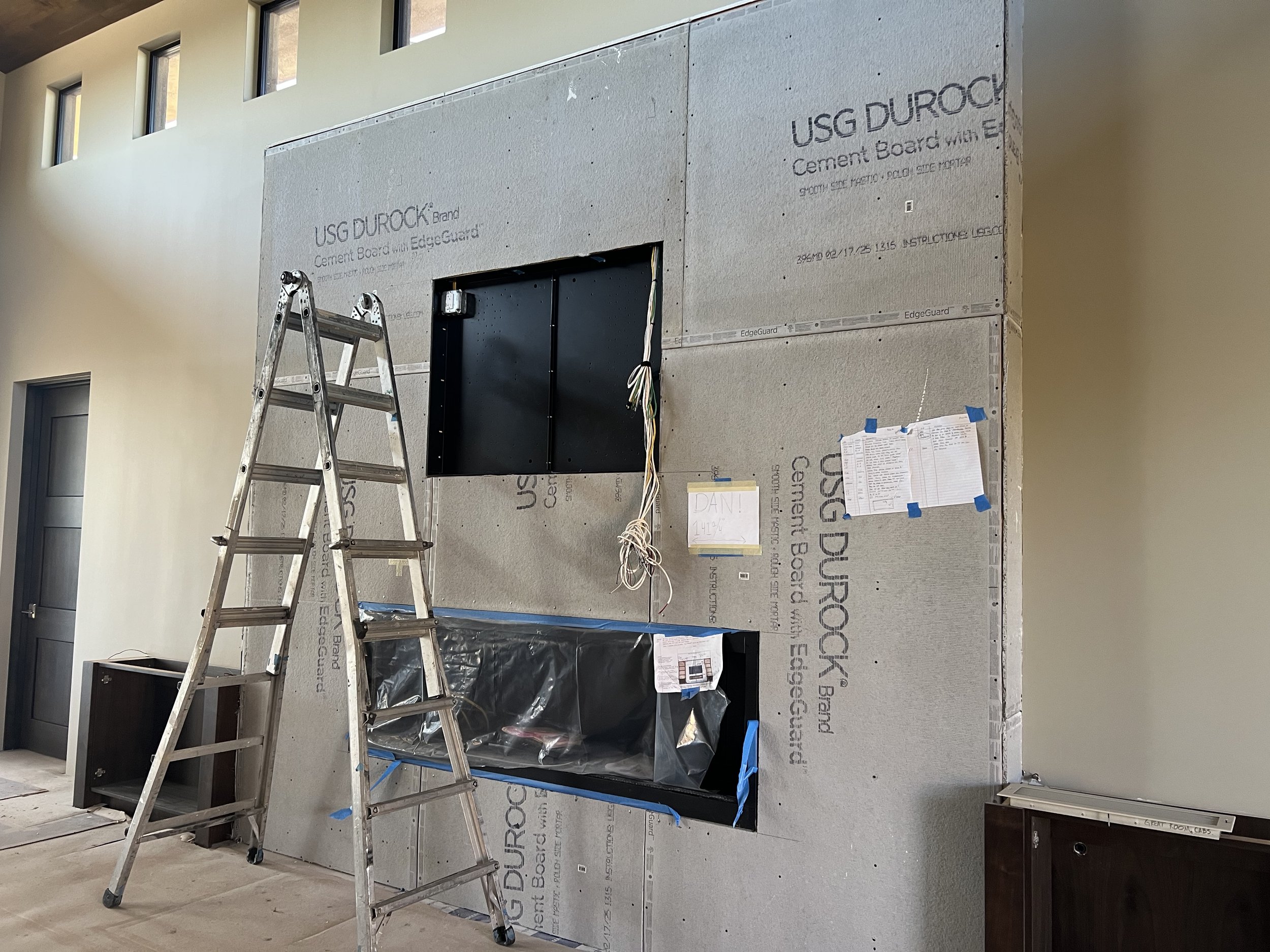 Interior view of a building under construction with an unfinished wall covered with USG Durock cement board, a metal ladder, and construction materials and papers taped to the wall.