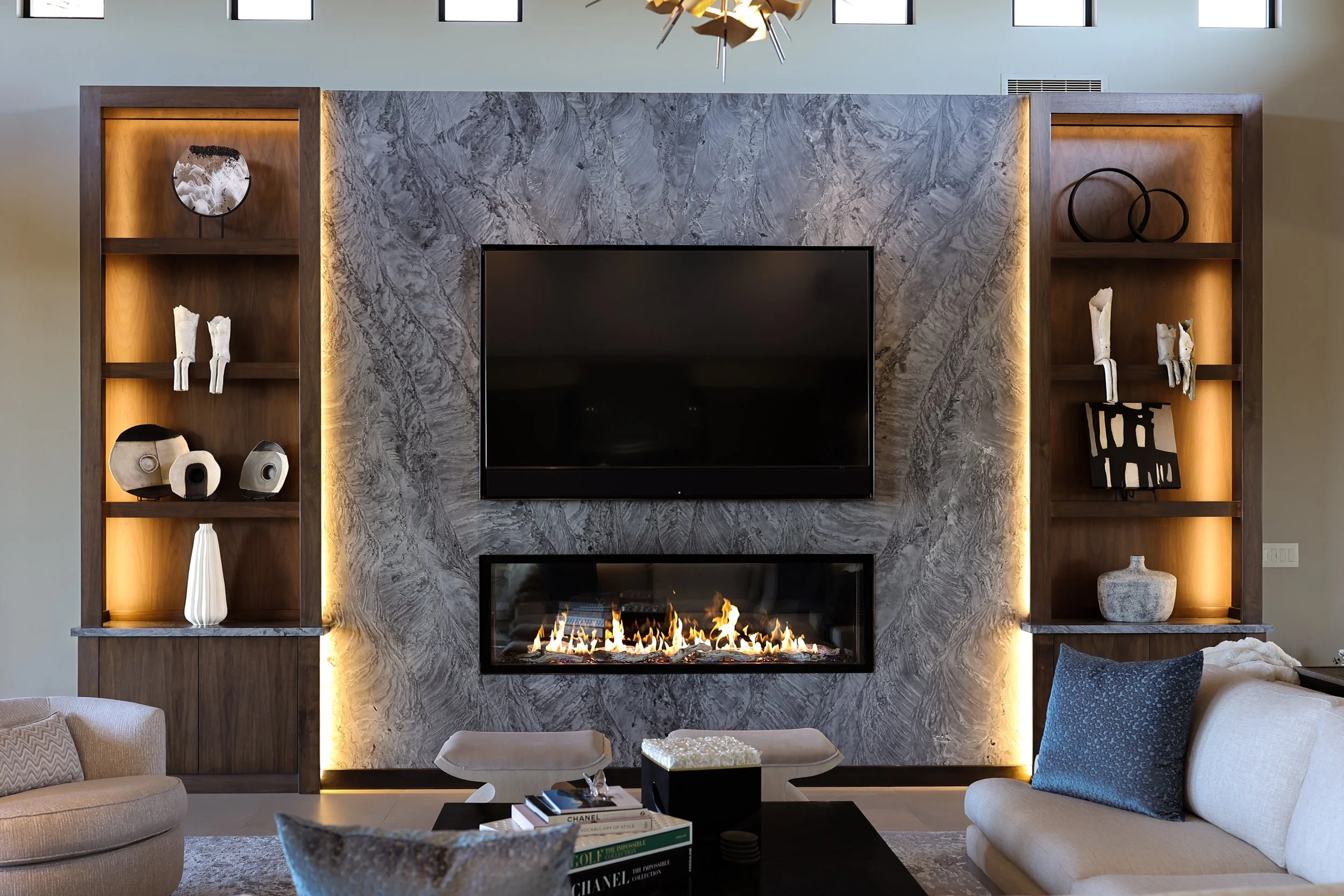 Modern living room with a stone fireplace, wall-mounted TV, and built-in wooden shelves decorated with art pieces and sculptures. Light enters through high windows.