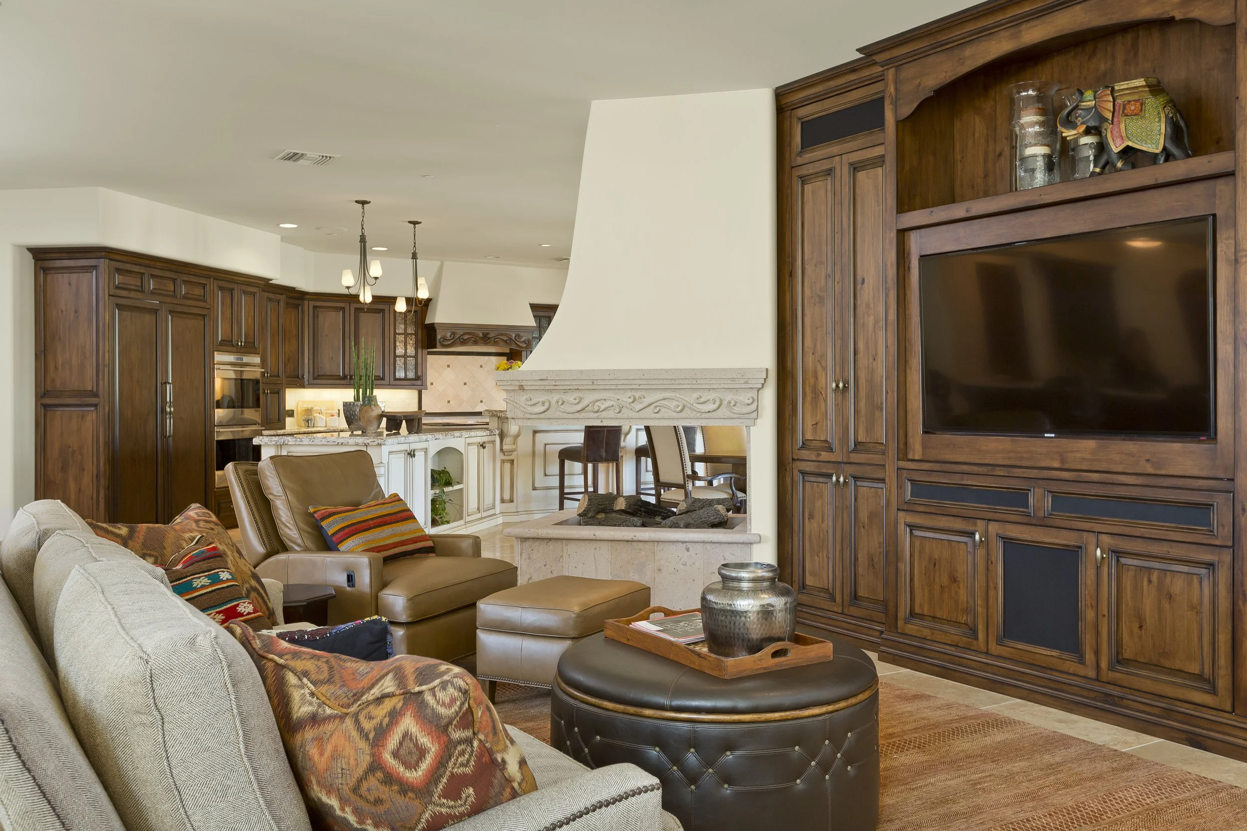 Living room with beige sofa, colorful patterned pillows, brown leather ottoman, and dark wood entertainment center with a flat-screen TV, decorated with glass vases and a wooden elephant sculpture, and a fireplace with dark stones.
