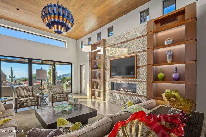 Living room with large windows, modern furniture, a flat-screen TV, decorative shelving with vases, and a wooden ceiling.