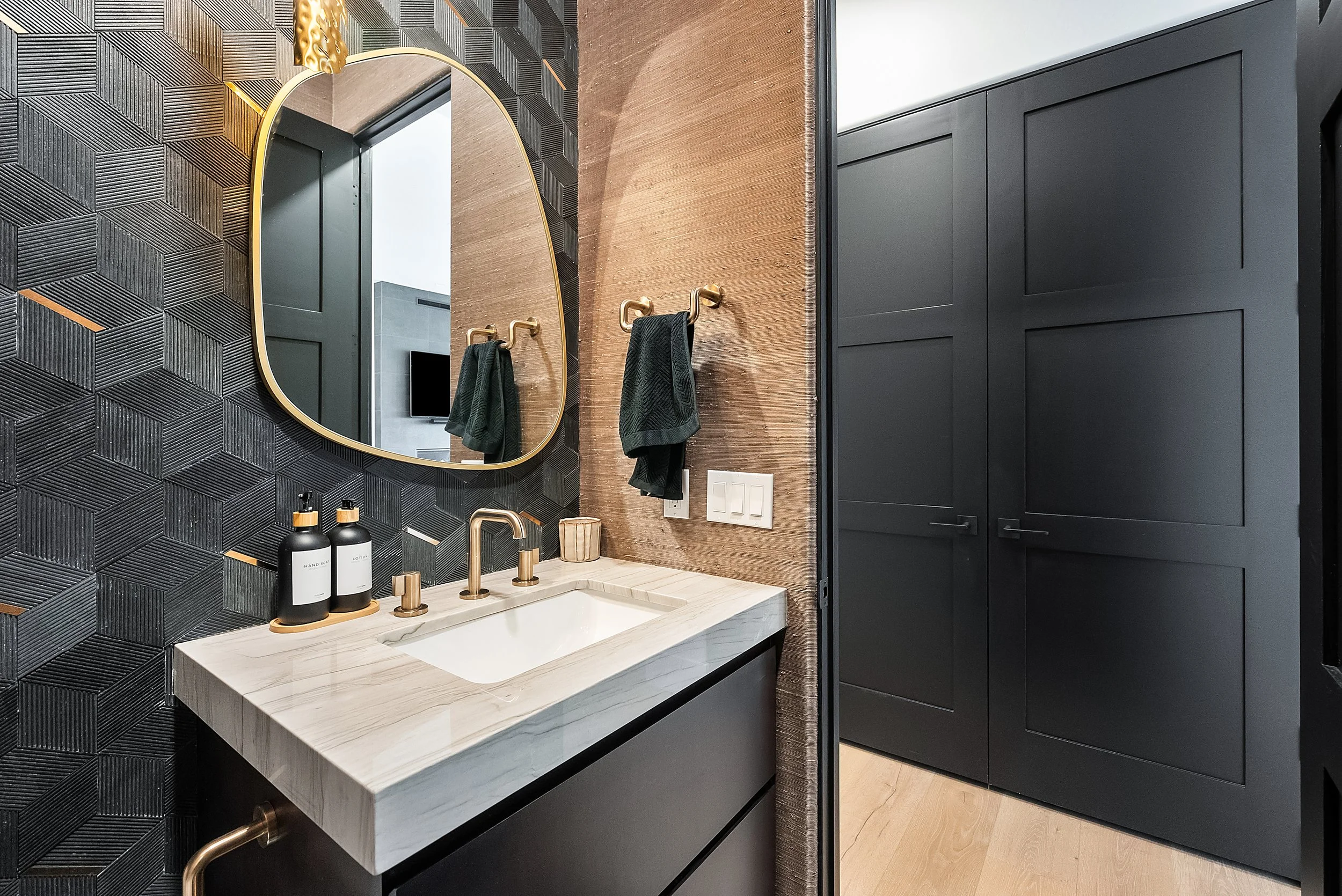 Modern bathroom vanity with a white marble countertop, a rectangular sink, and a gold faucet. Behind the sink is a black textured tile wall, a large oval mirror with a gold frame, and black cabinets. Bottles of hand soap and lotion are on the counter