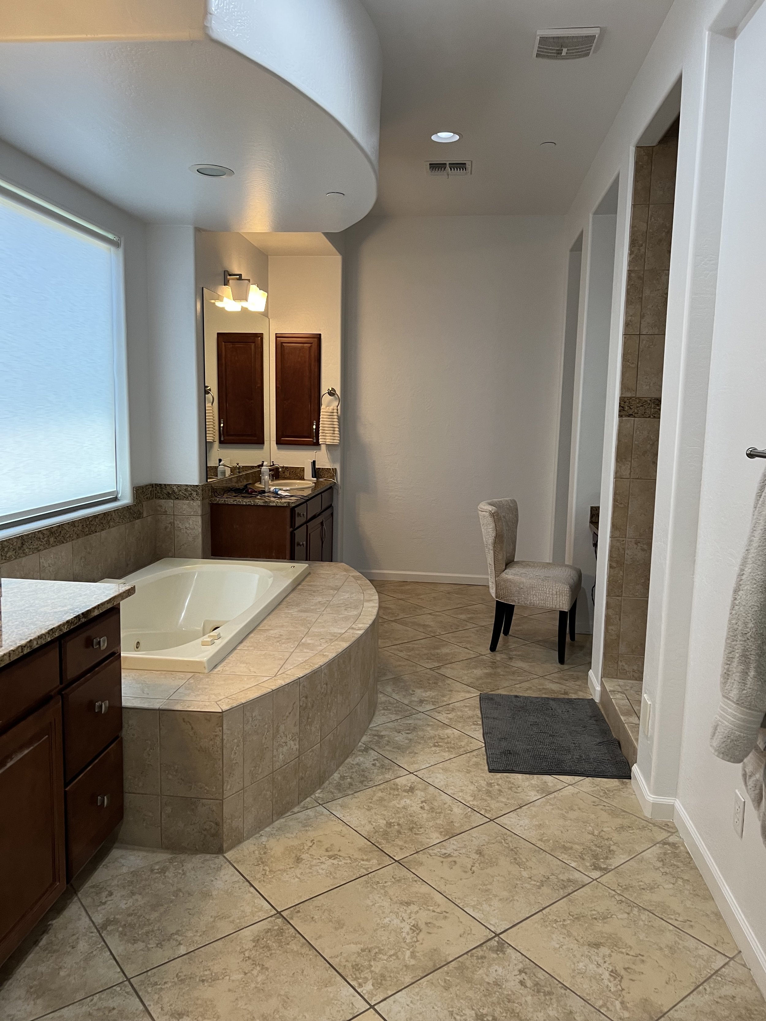 Bathroom with a bathtub, wooden cabinet with a mirror, beige tiled floors and walls, a window with blinds, a chair, a towel rack, and various toiletries on the sink.