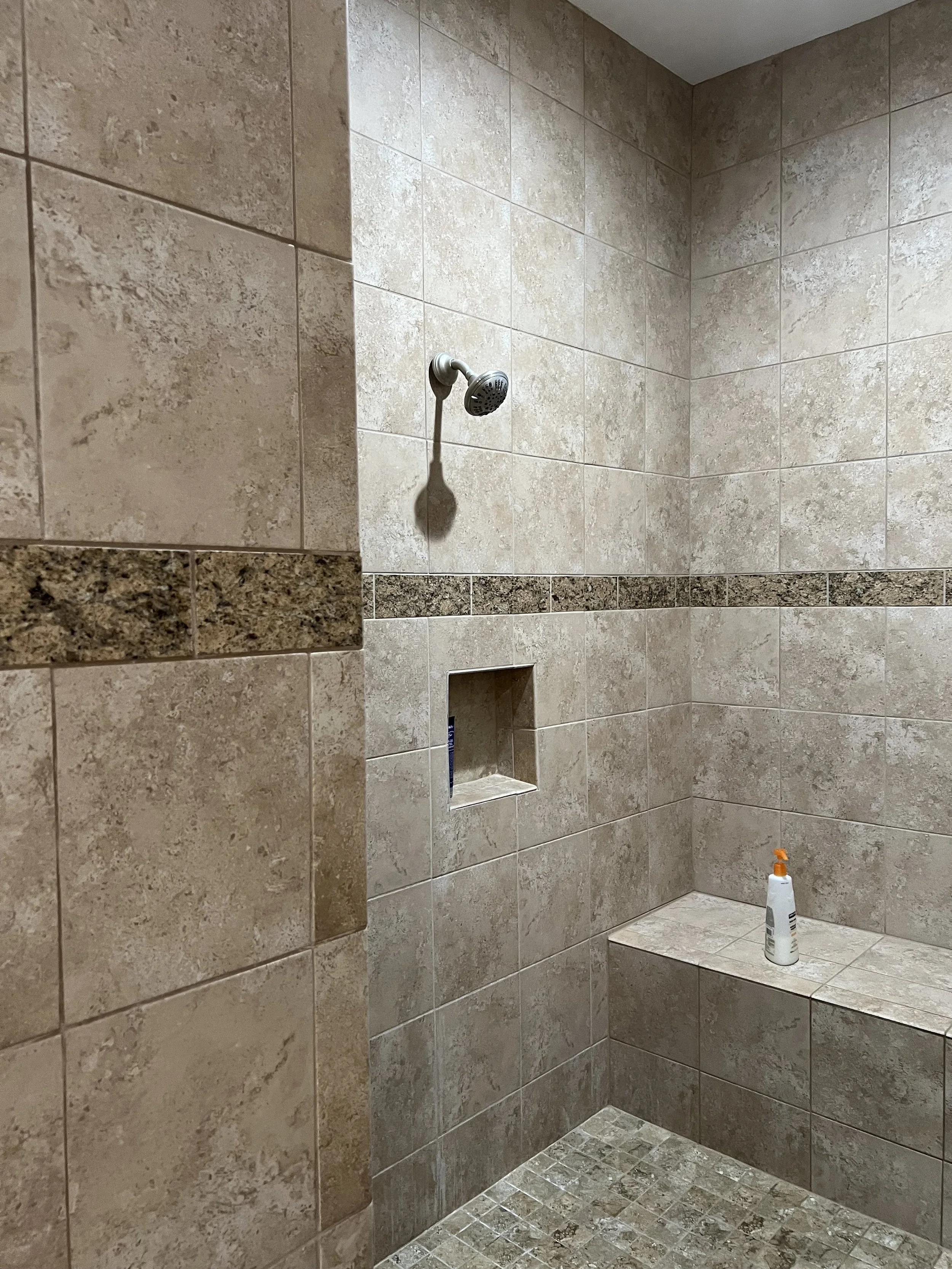 A walk-in shower with beige and brown tiled walls, a built-in niche for toiletries, and a bottle of lotion on a ledge.