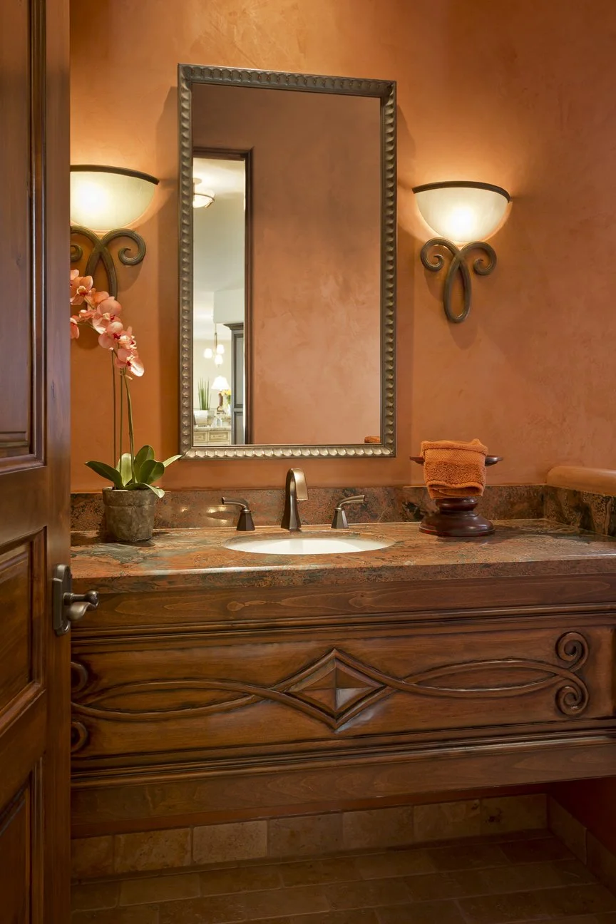 A bathroom sink with a wooden vanity and a granite countertop, a mirror, wall sconces, a potted orchid, and a folded orange towel.