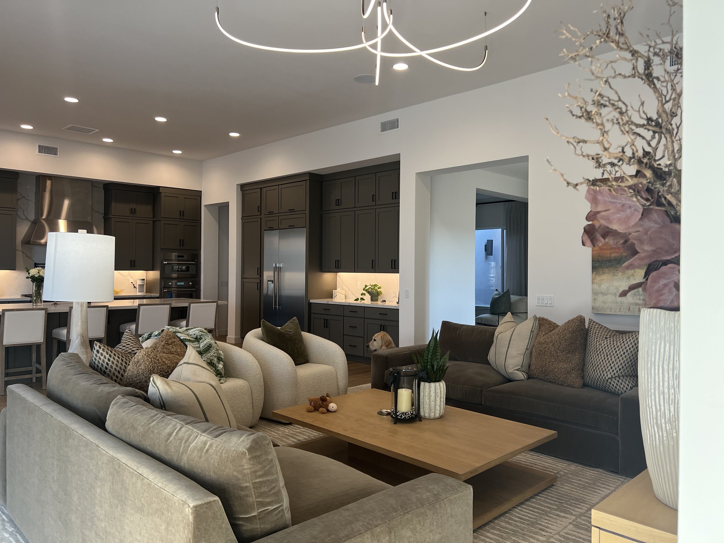 Living room with beige couches, a brown couch, and decorative pillows. Coffee table with candles and stuffed animals. Modern lighting and kitchen area in background.