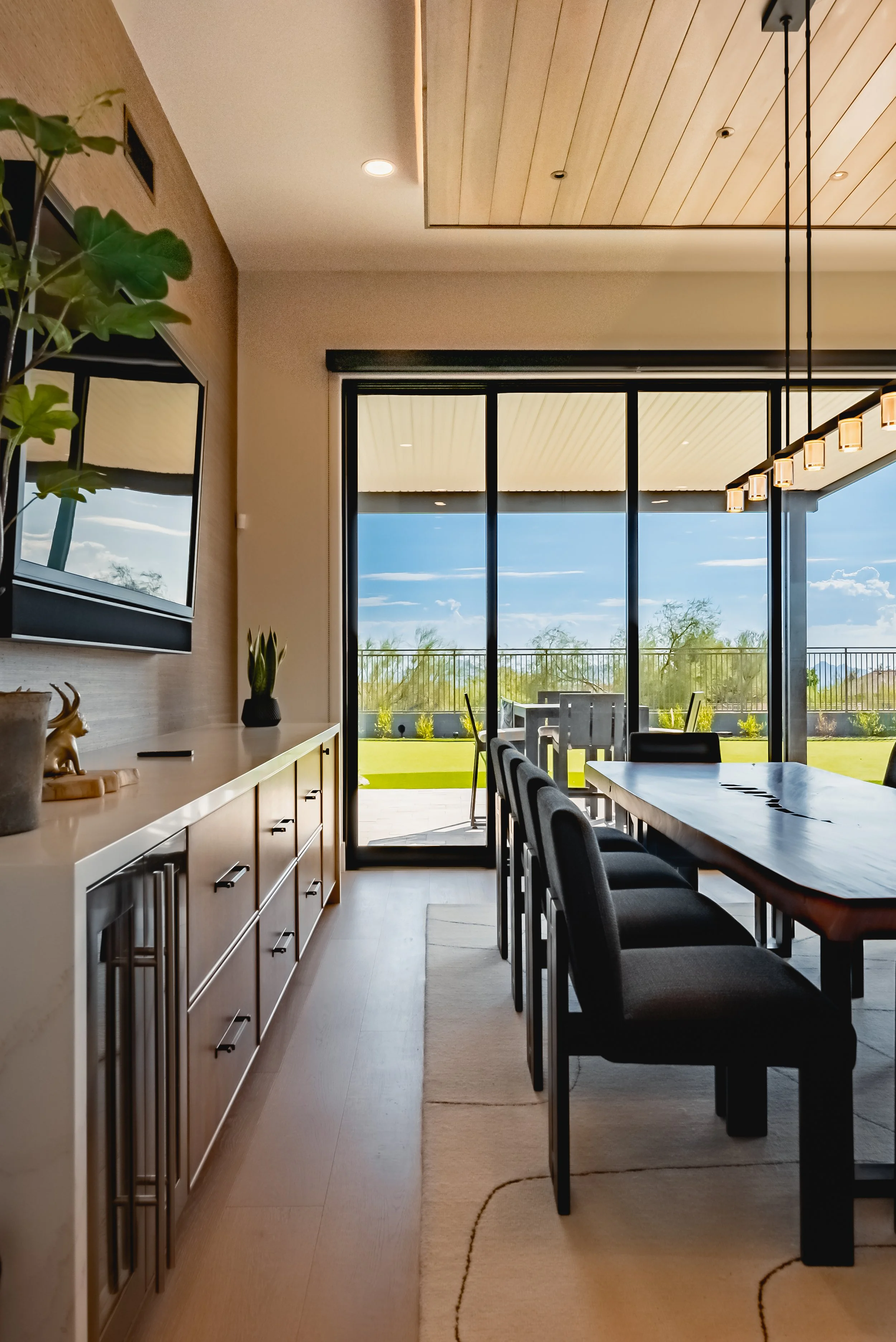 A spacious dining room with a long wooden table with black chairs, large floor-to-ceiling glass windows, a view of trees and a blue sky outside, a modern pendant light, and a sideboard with decorative items and plants.