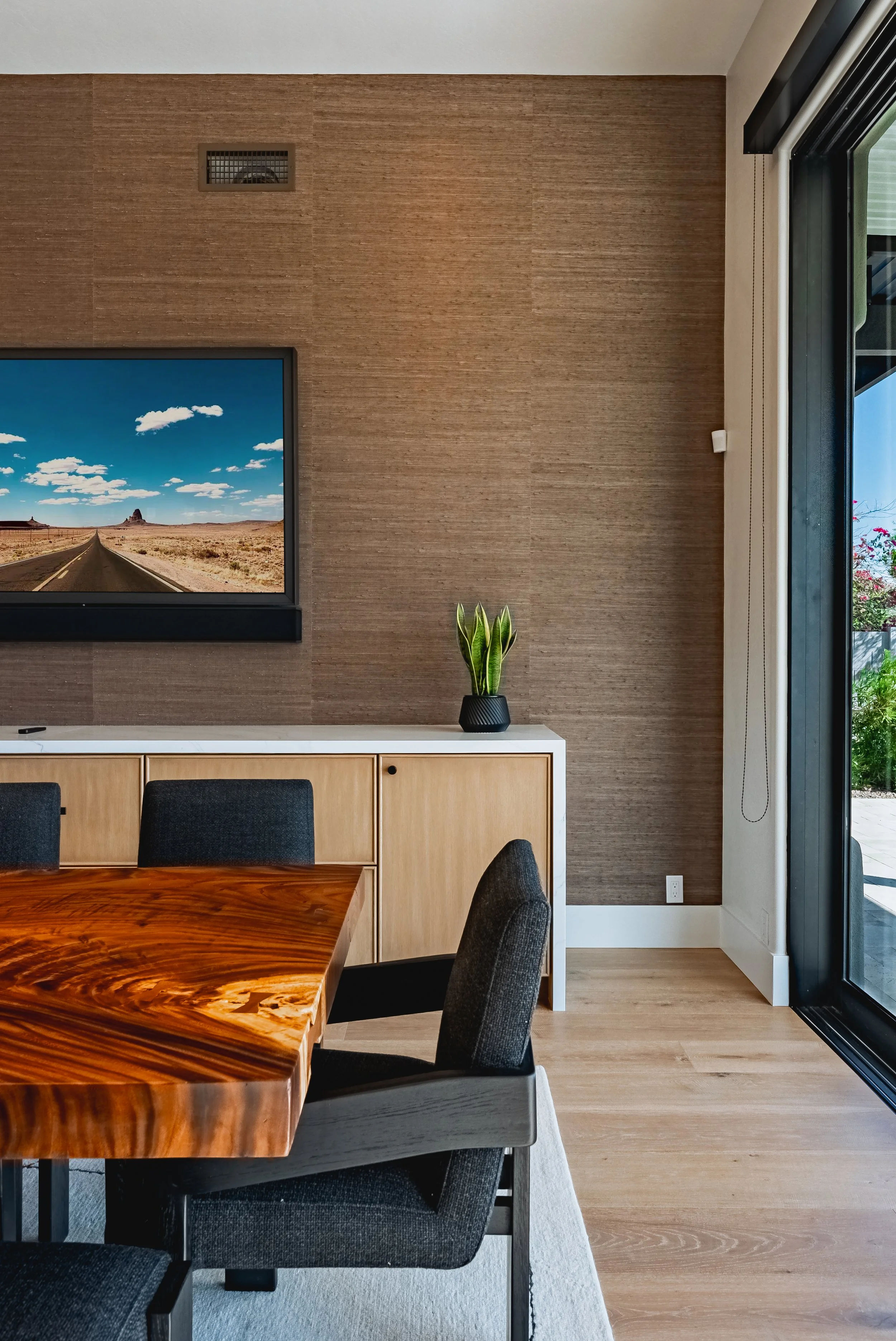 A modern dining area with a circular wooden table, black upholstered chairs, a wooden sideboard with a potted plant, a large flat-screen TV, a textured brown wall, and a glass sliding door leading outside.