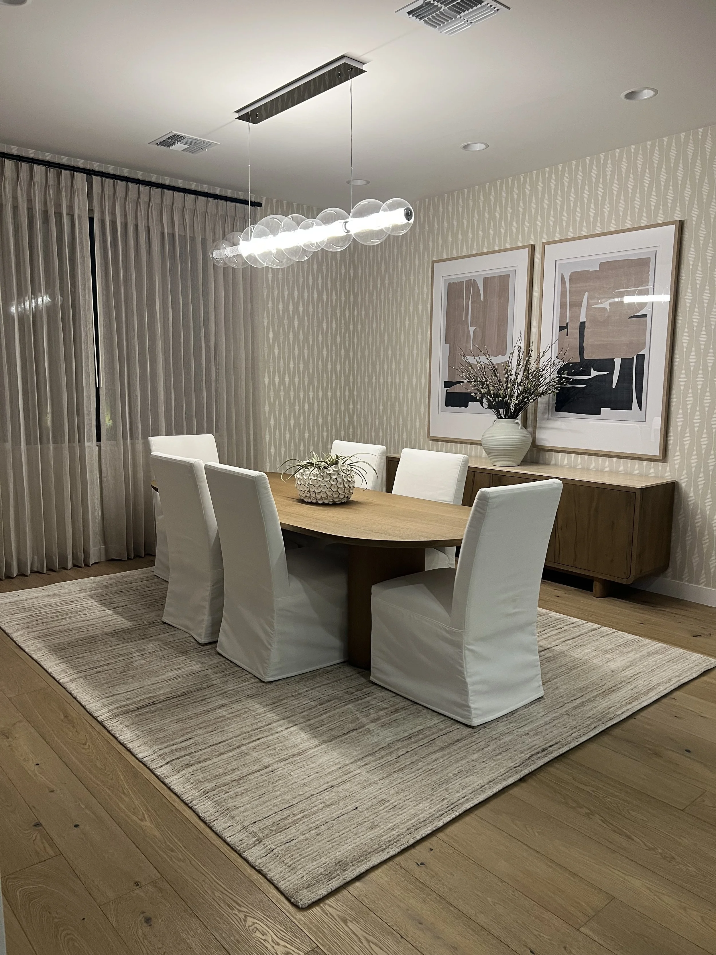 A modern dining room with a wooden table and six white slipcovered chairs on a large cream-colored area rug. The wall has framed abstract artwork and a tall vase of branches on a wooden sideboard. A contemporary chandelier with glass globes hangs abo