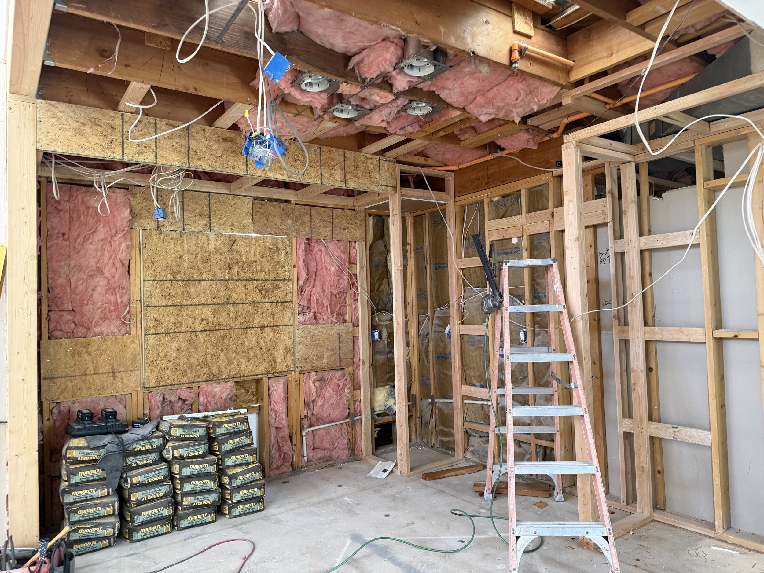 Interior of a house under construction or renovation showing exposed wooden framing, pink insulation, wiring, and electrical outlets, with a step ladder and construction tools.