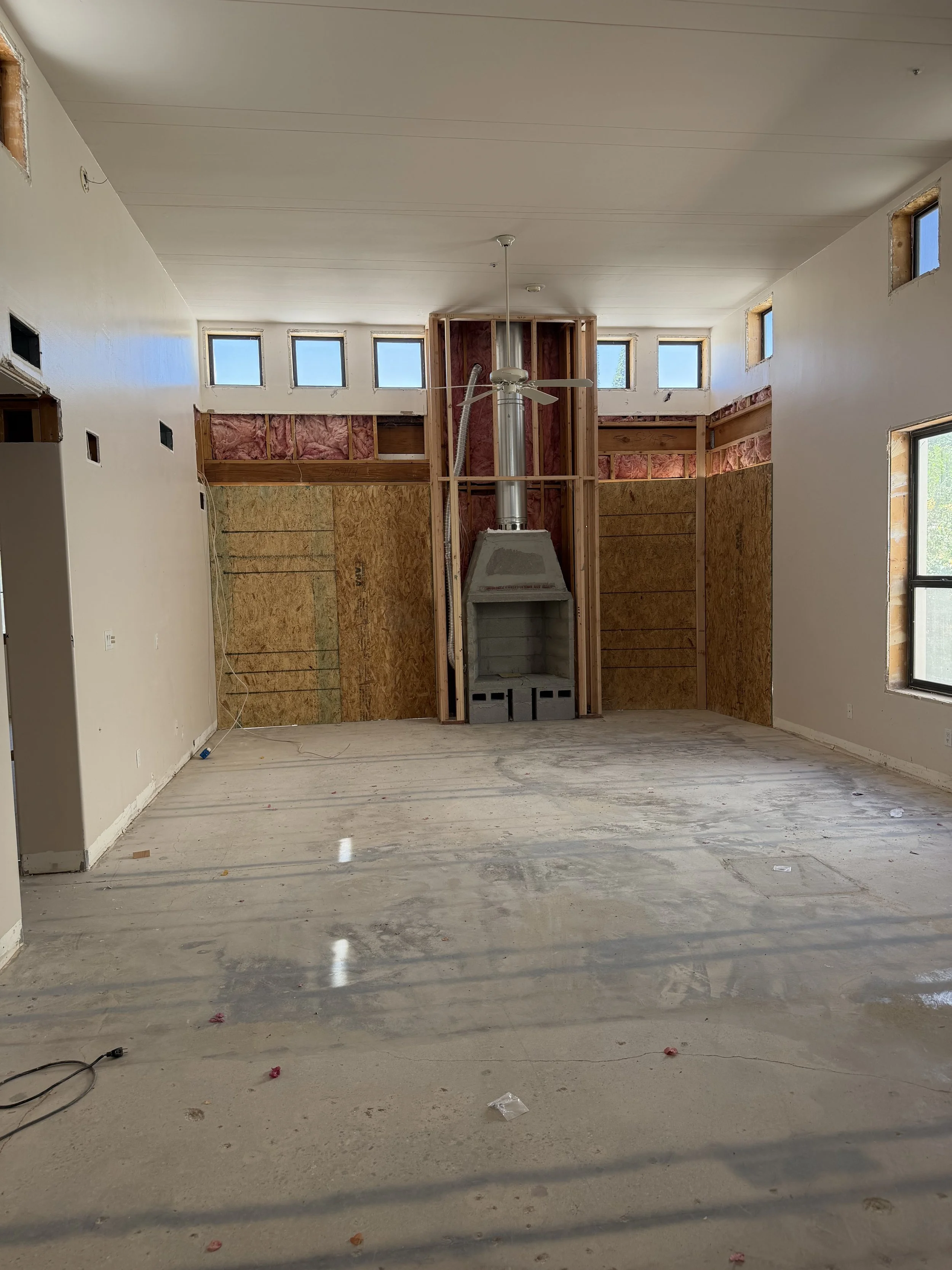 An unfinished room under construction with a centered fireplace and a ceiling fan, with multiple small windows along the upper part of the walls, and construction materials and debris on the floor.