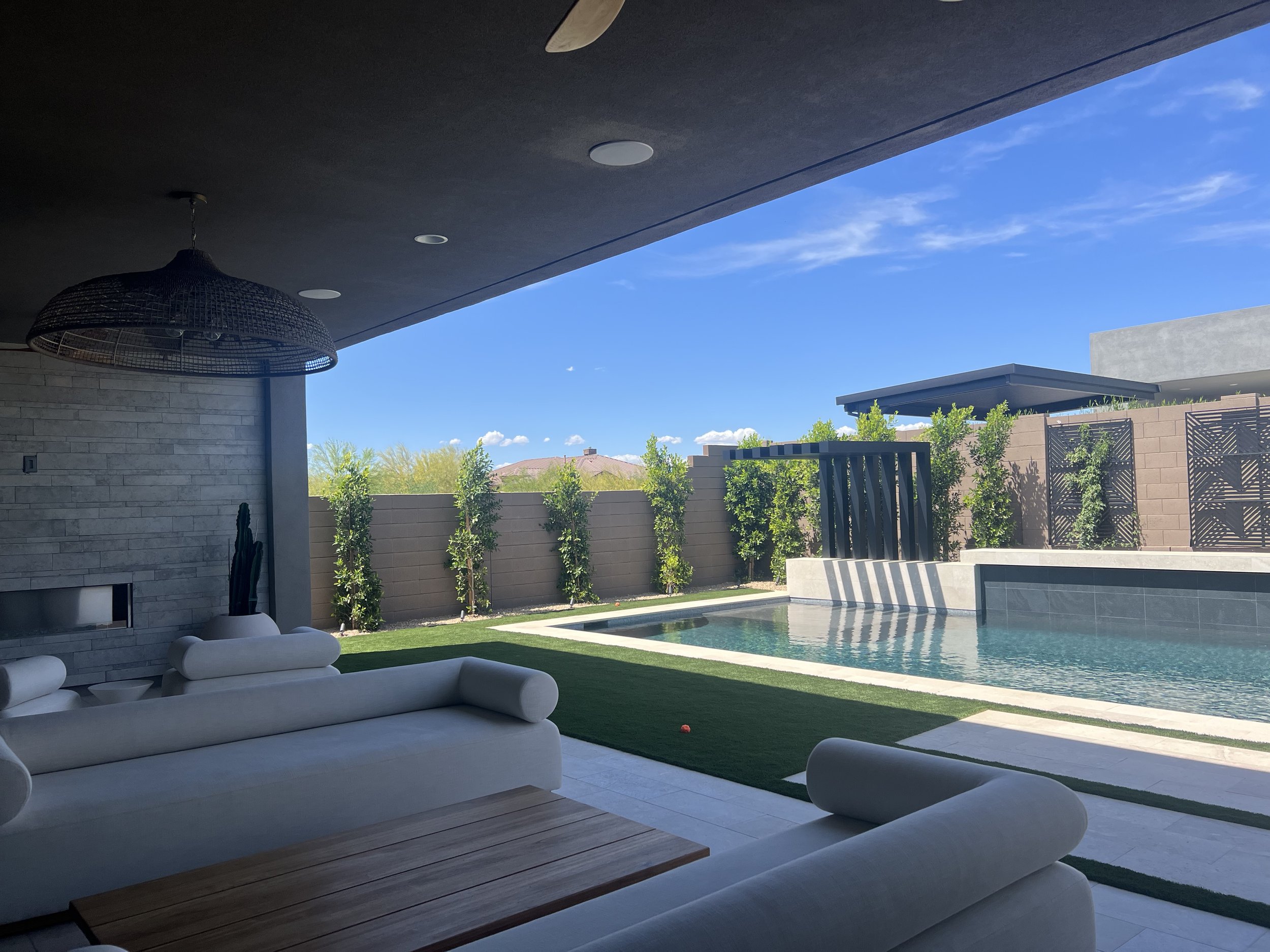 Modern outdoor patio area with white lounge chairs, a swimming pool, green lawn, and a privacy fence with tall plants, under a clear blue sky.