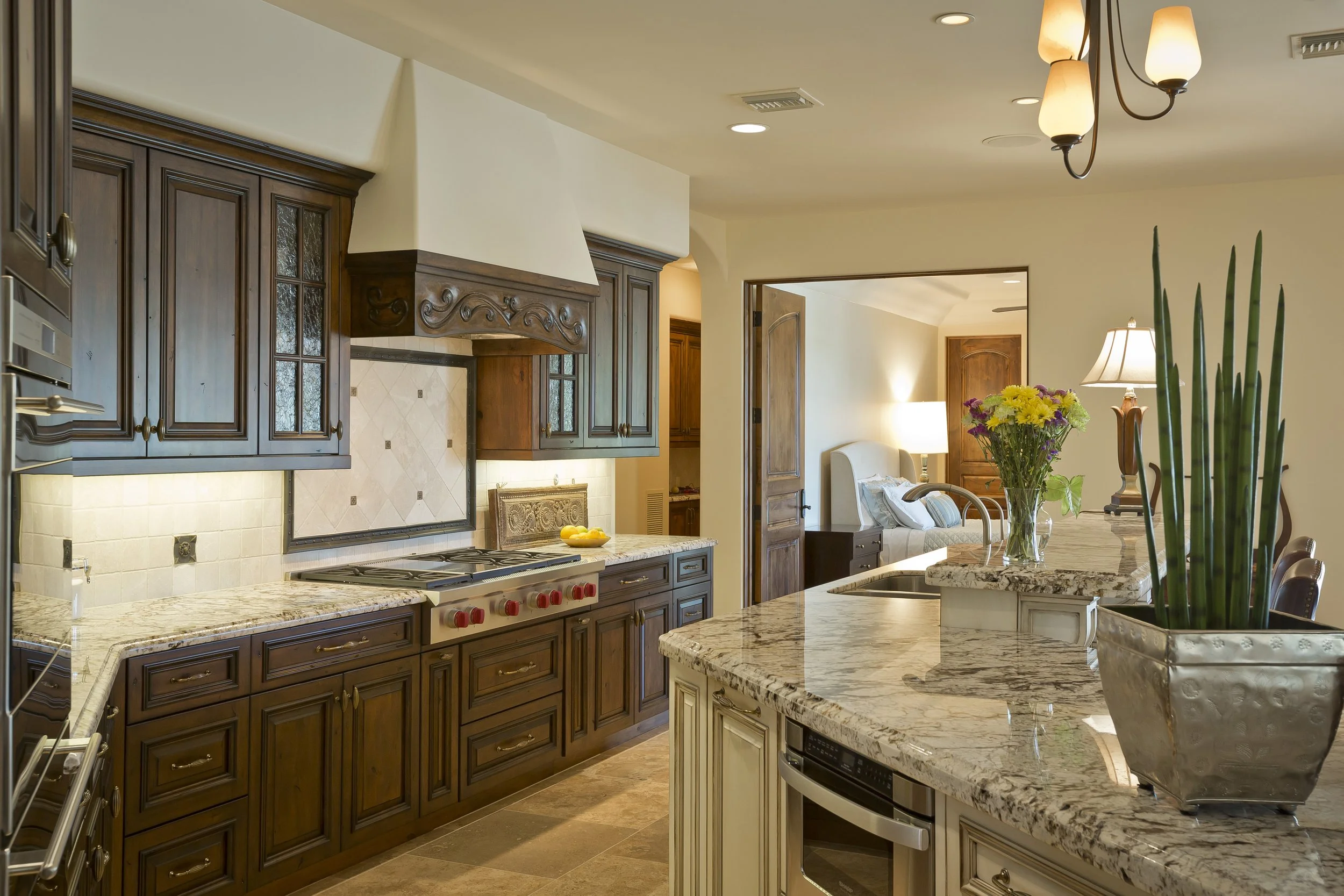 A kitchen with dark wooden cabinets, a granite countertop island, and a tiled backsplash. There is a stove with red knobs and a hood above it. In the background, a living area with a sofa, a table lamp, and a bouquet of yellow and purple flowers on t