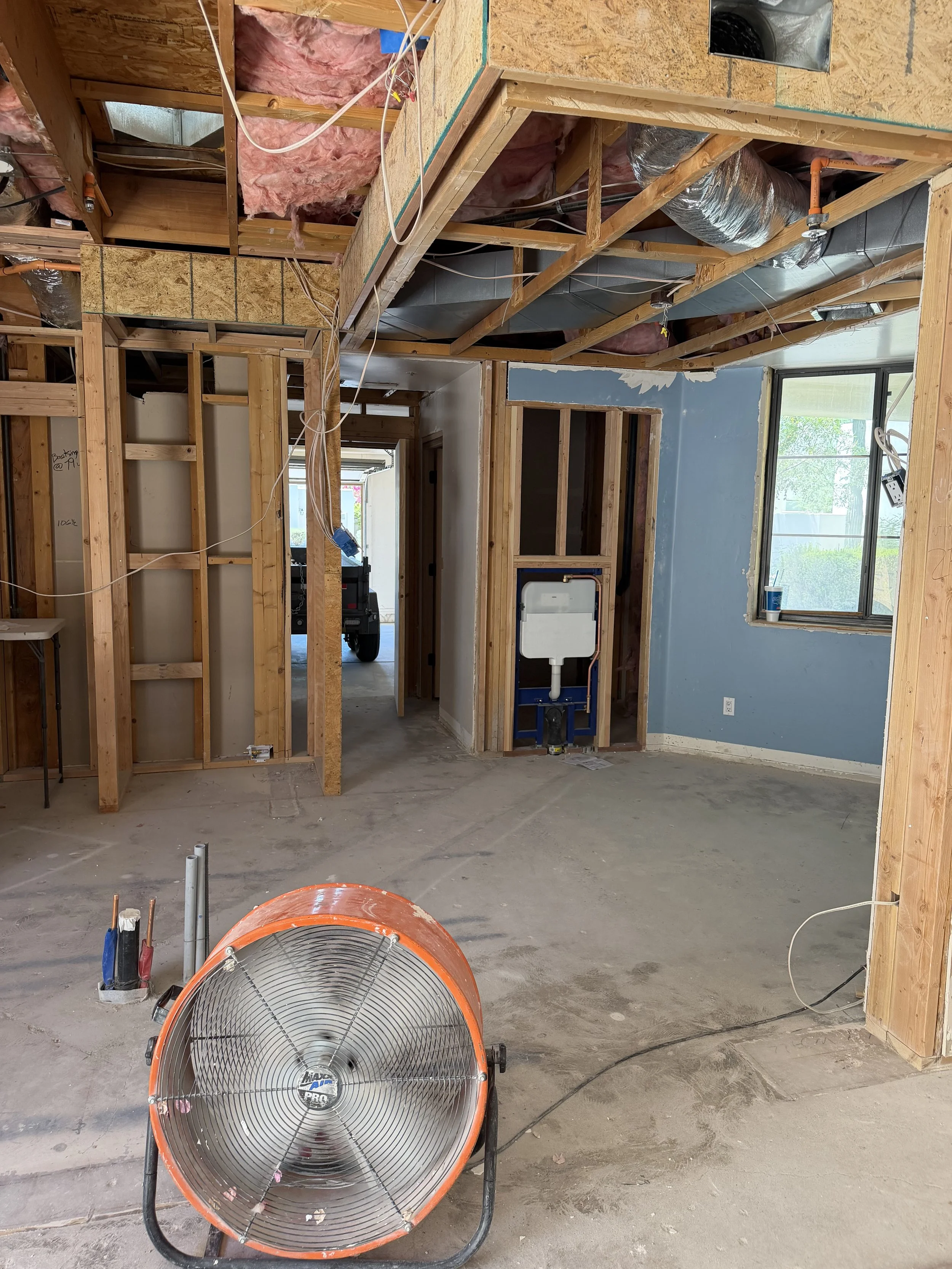 An unfinished room under construction with exposed framing, insulation, and ductwork; a large industrial fan positioned on the concrete floor, and a window allowing natural light in.