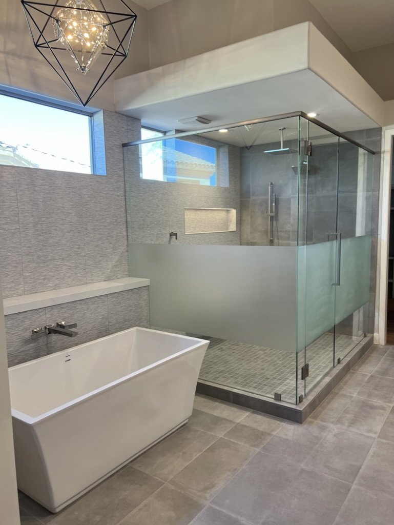Modern bathroom featuring a freestanding bathtub and a glass-enclosed shower with gray tile walls and flooring, two high windows providing natural light, and a contemporary geometric pendant light fixture.
