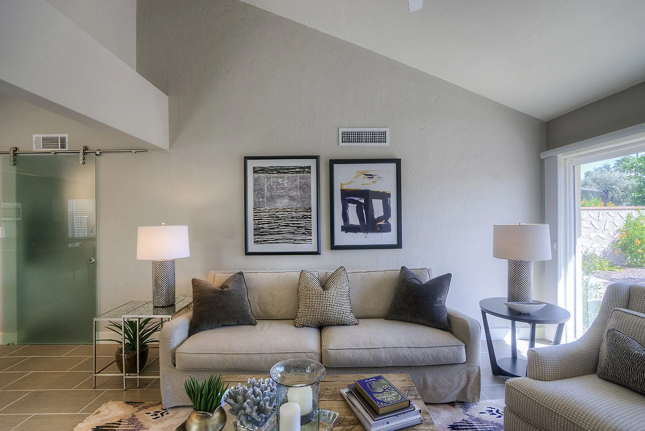 Living room with beige sofa, armchair, glass coffee table, side tables with lamps, wall art, and sliding glass door leading outside.