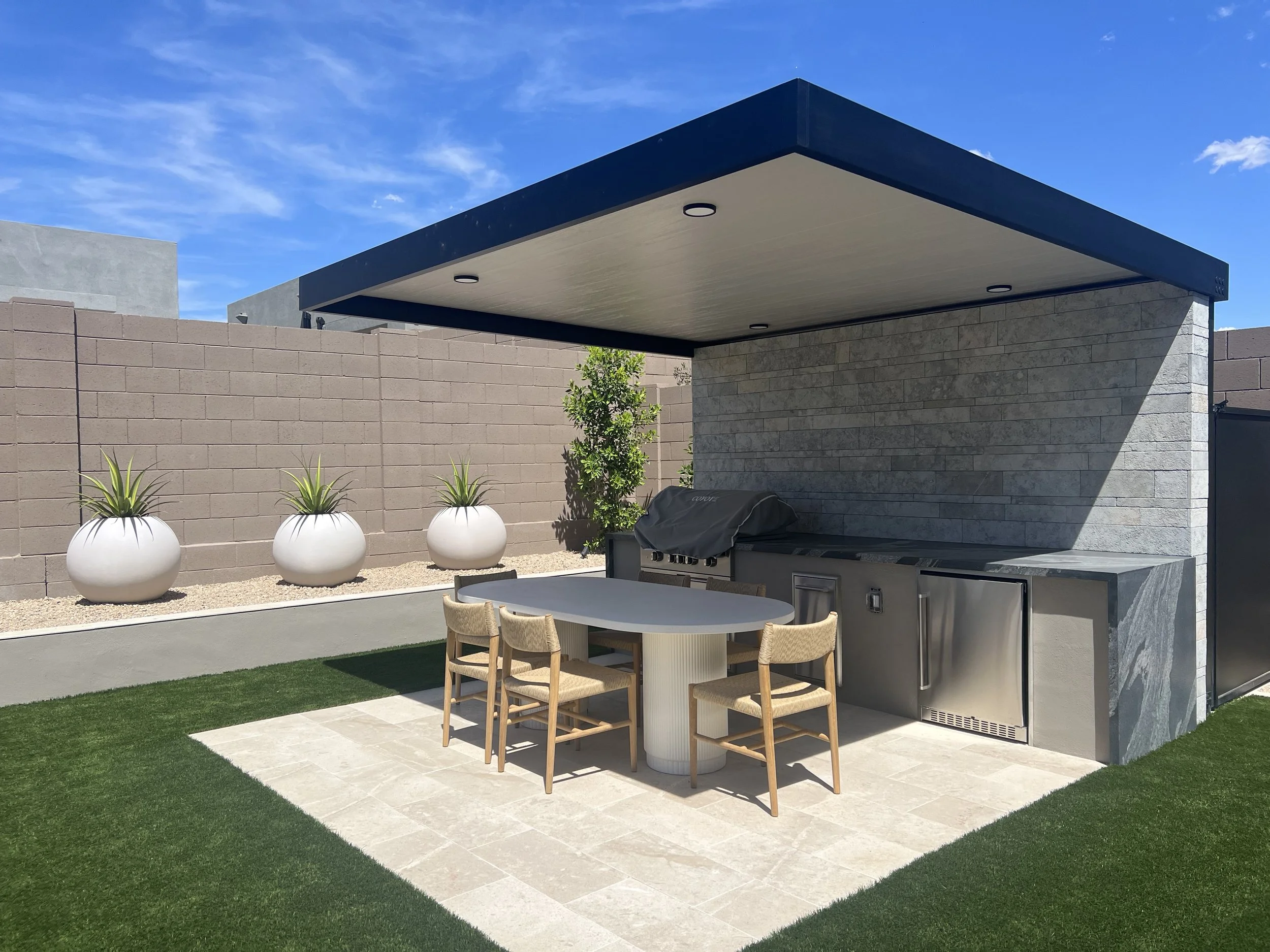 A modern outdoor patio area with a covered seating and cooking space, featuring a table with chairs, potted plants, a brick wall, a grassy area, and a blue sky.