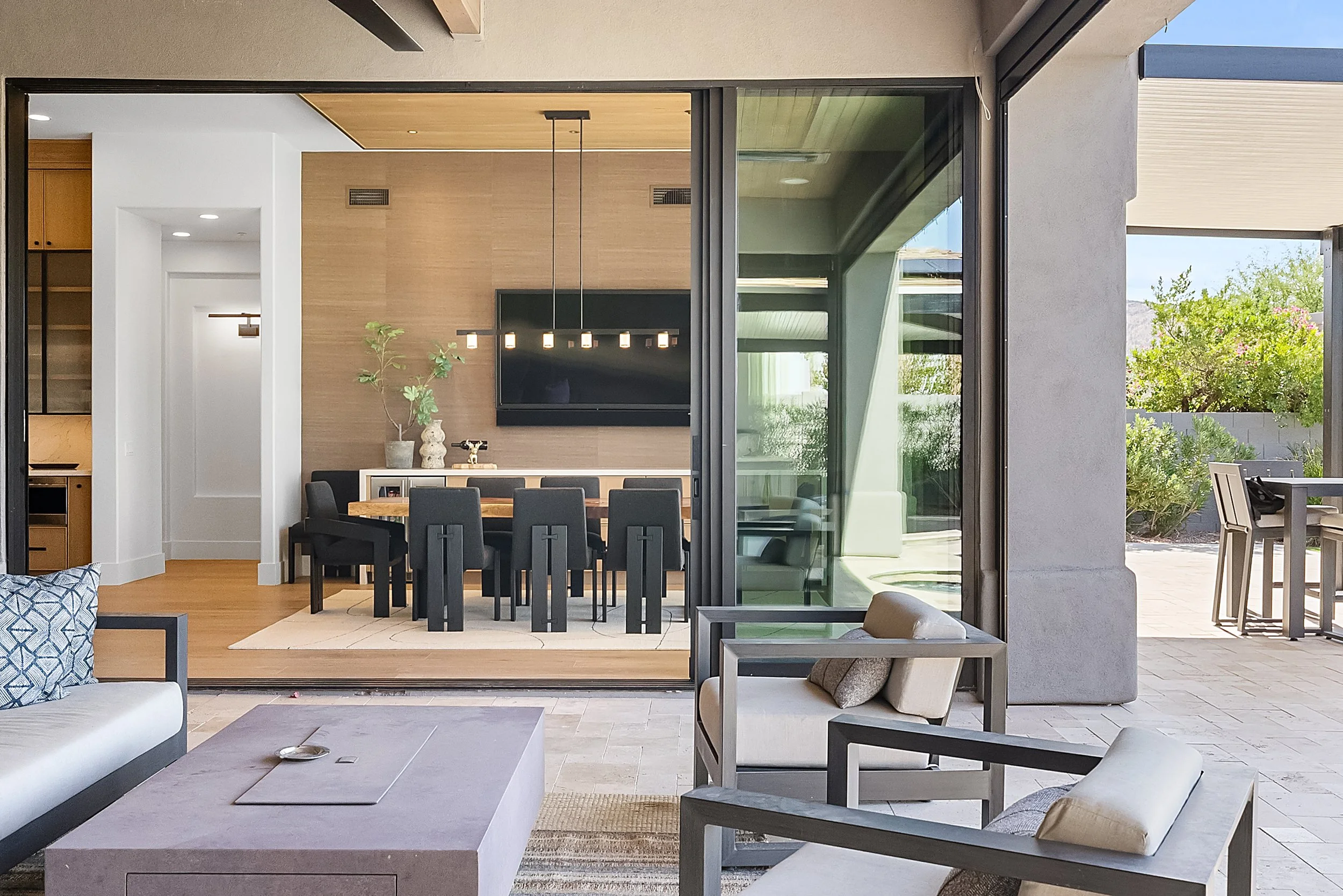 Modern indoor and outdoor living space with a dining area, seating, and a view of a backyard with greenery.