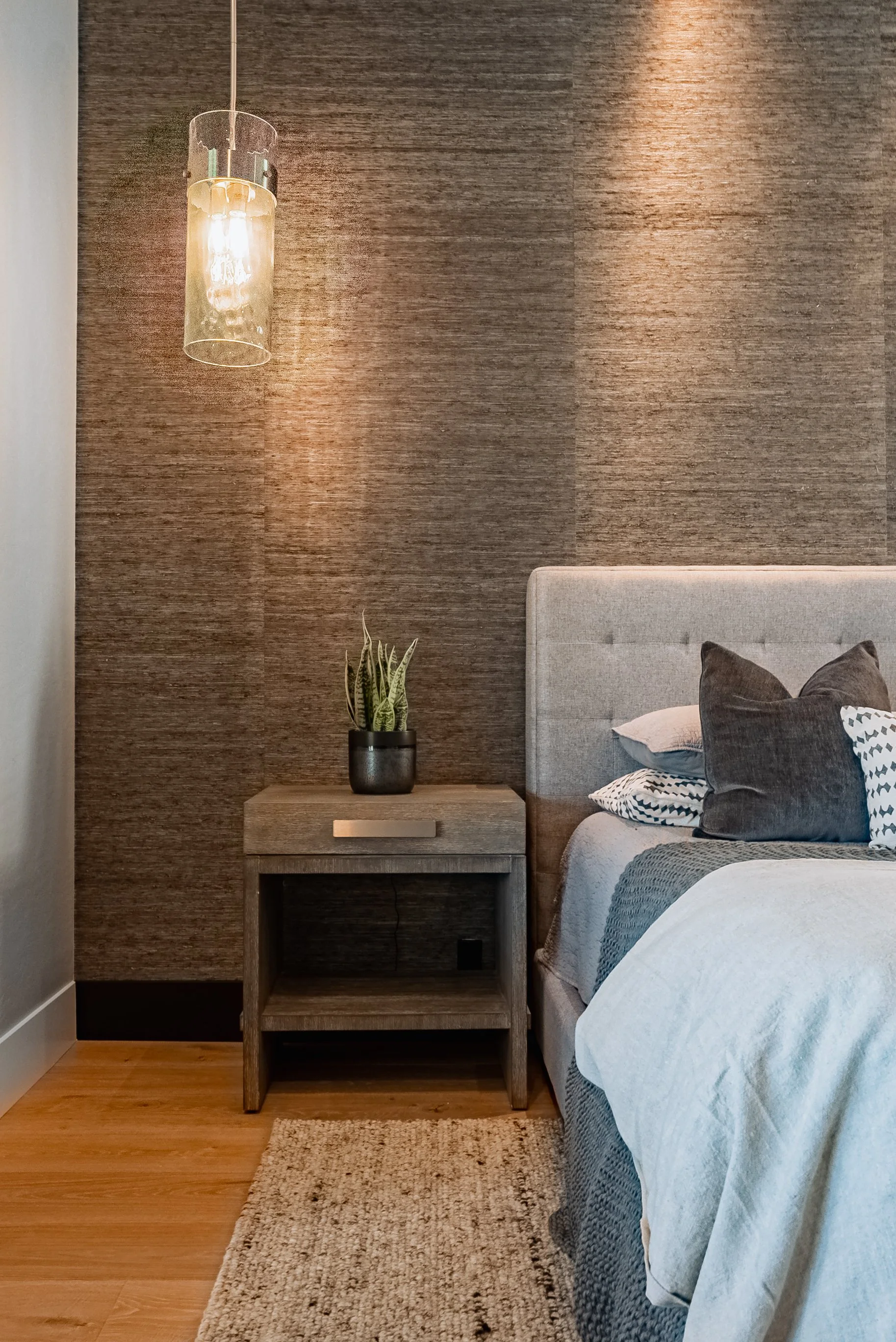 A bedroom with a bed, gray and white bedding, and multiple pillows. There is a wooden nightstand with a black plant pot containing a green plant. A textured beige rug is on the wooden floor. The wall behind the bed has brown textured wallpaper, and a