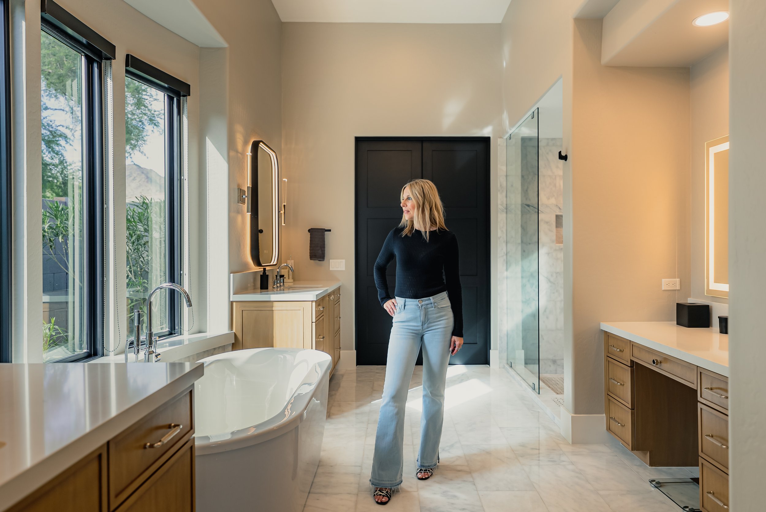 A woman in light blue jeans and a black sweater stands in a modern bathroom with large windows, a bathtub, and cabinetry, looking to the side.