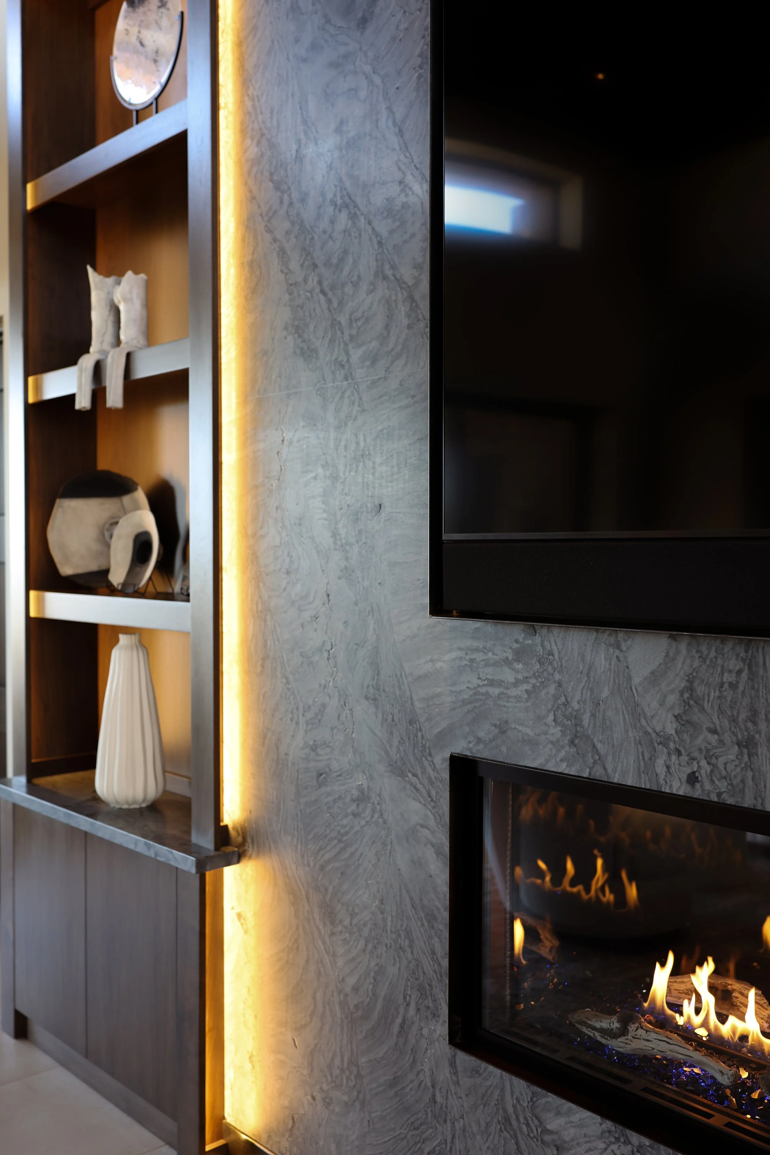 Close-up view of a modern living room with a fireplace, marble wall, wooden shelf with decorative items, and warm lighting.