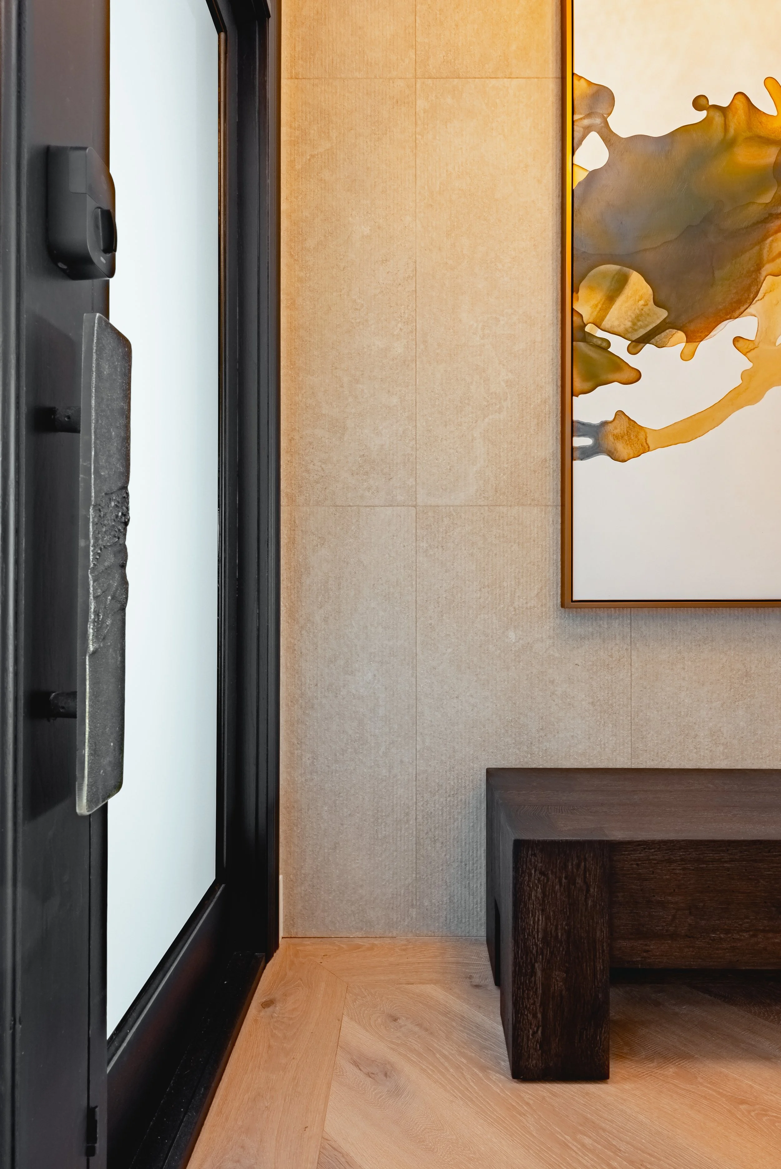 Interior view of a room showcasing a large, white projection screen with a black frame, a dark wood bench, and a decorative wall with abstract art in earth tones. The floor is light wood, and the wall behind has a textured beige surface.